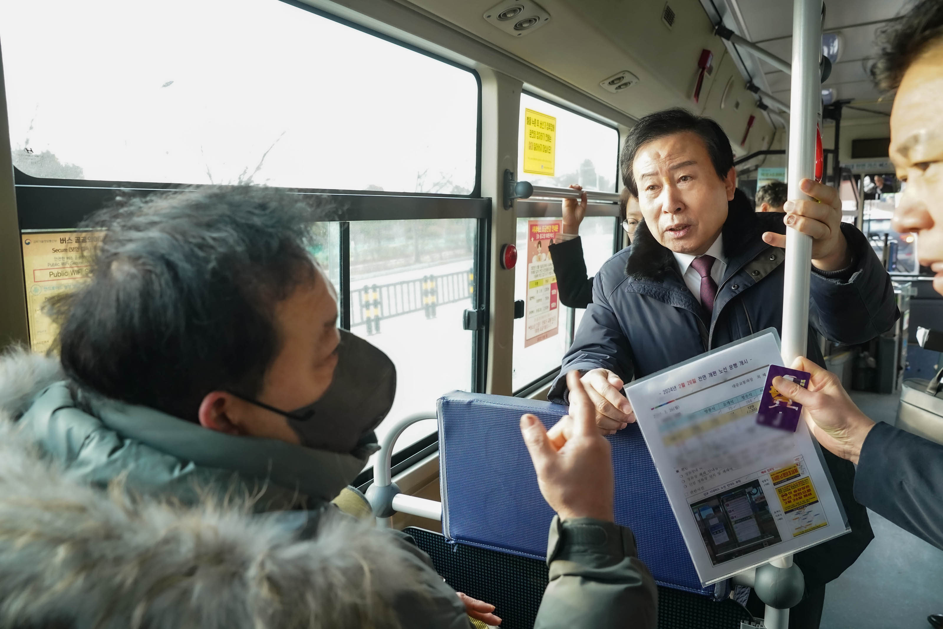 버스 좌석에 앉아 짙은 회색 마스크를 착영하고 카키색 패딩을 입은 시민과 이야기를 나누는 박홍률 시장과 의원. 의원은 옆모슾만 보이고, 손에는 노선 개편과 관련된 자료와 보라색 교통카드를 쥐고 있다.