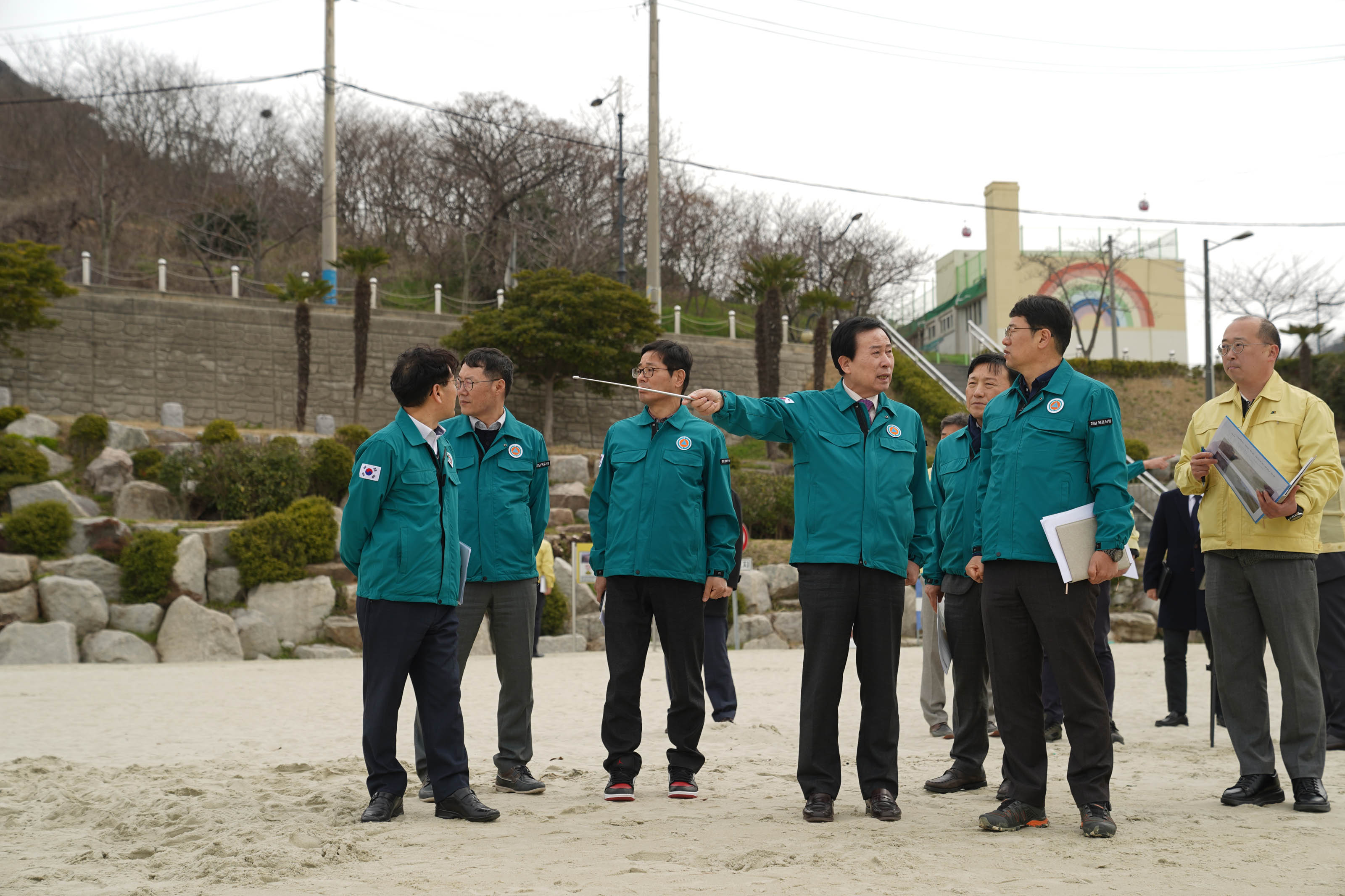 모래로 된 바닥에 박홍률 시장과 직원들이 7명 이상 서있고, 박홍률 시장은 지침봉으로 무언가를 가르키며 이야기 하고 있다.