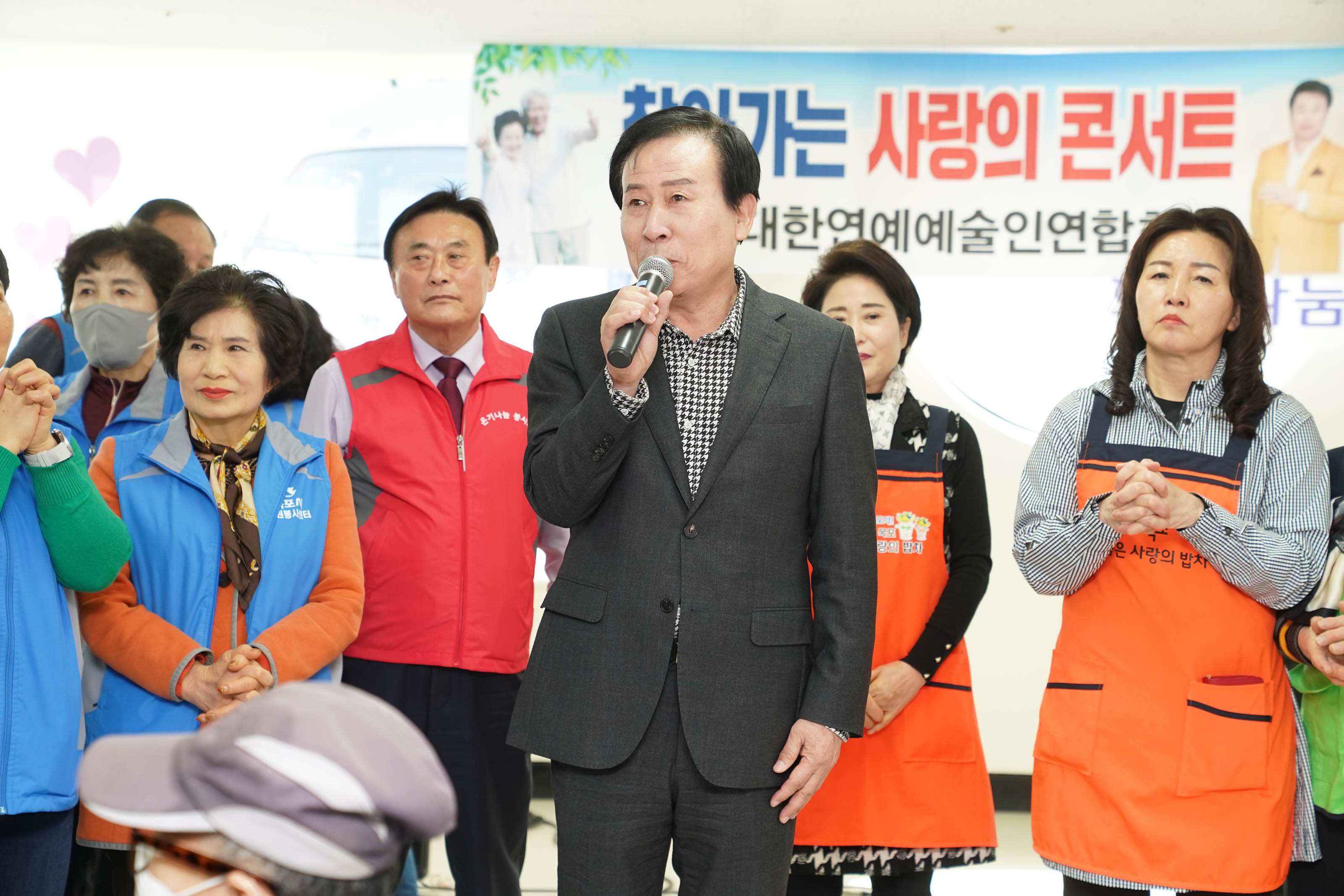 마이크를 쥐고 얘기하고 있는 박홍률 시장 뒤로 빨강, 파랑의 봉사 유니폼을 입은 5여명의 사람들과 주황색의 밥차 앞치마를 두른 두명의 여성이 서있다.