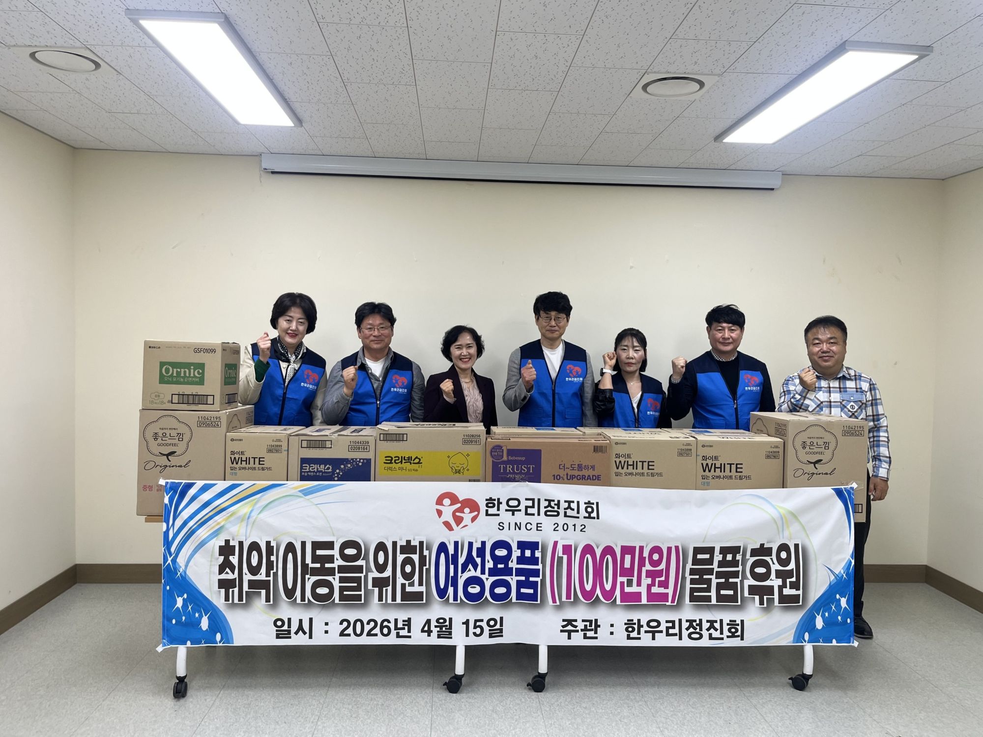 한우리정진회 SINCE 2012 취약 아동을 위한 여성용품(100만원)물품 후원 일시:2025년 4월 15일, 주관: 한우리정진회 문구가 적힌 현수막이 보이고, 테이블에 여성용품 박스 약 10개가 보인다. 박스 뒤로 화이팅을 하며 웃고 있는 7명의 회원들이 있다.