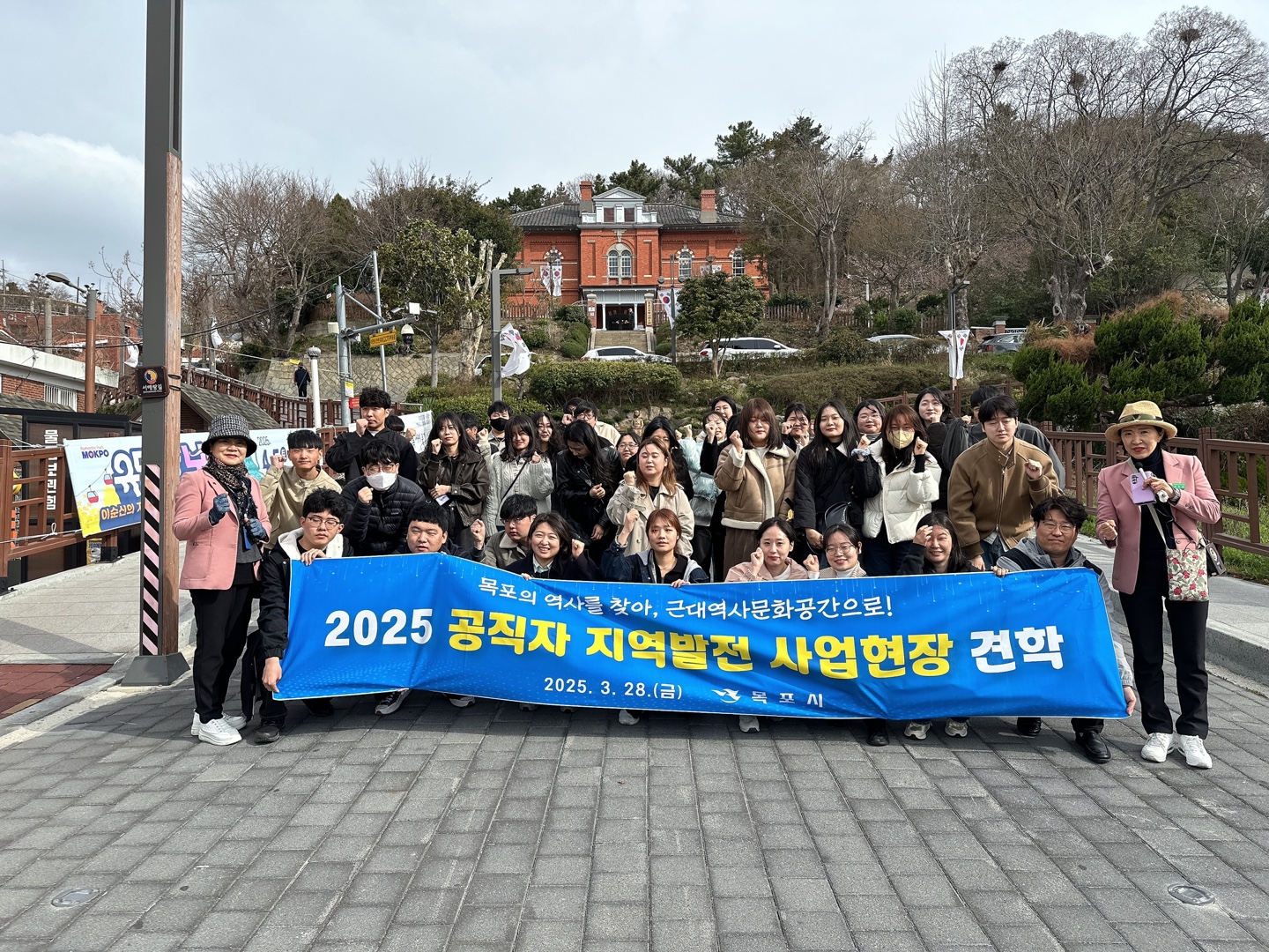 2025 공직자 지역발전 사업현장 견학에 참여한 공무원들이 파란색 현수막을 들고 단체 사진을 찍고 있는 모습. 현수막에는 '목포의 역사를 찾아, 근대역사문화공간으로! 2025.3.28(금)'이라는 문구가 적혀 있으며, 뒤편에는 붉은 벽돌 건물과 계단, 주변 나무들이 배경으로 어우러져 있다. 참가자들은 주먹을 들어 다짐의 포즈를 취하고 있다.