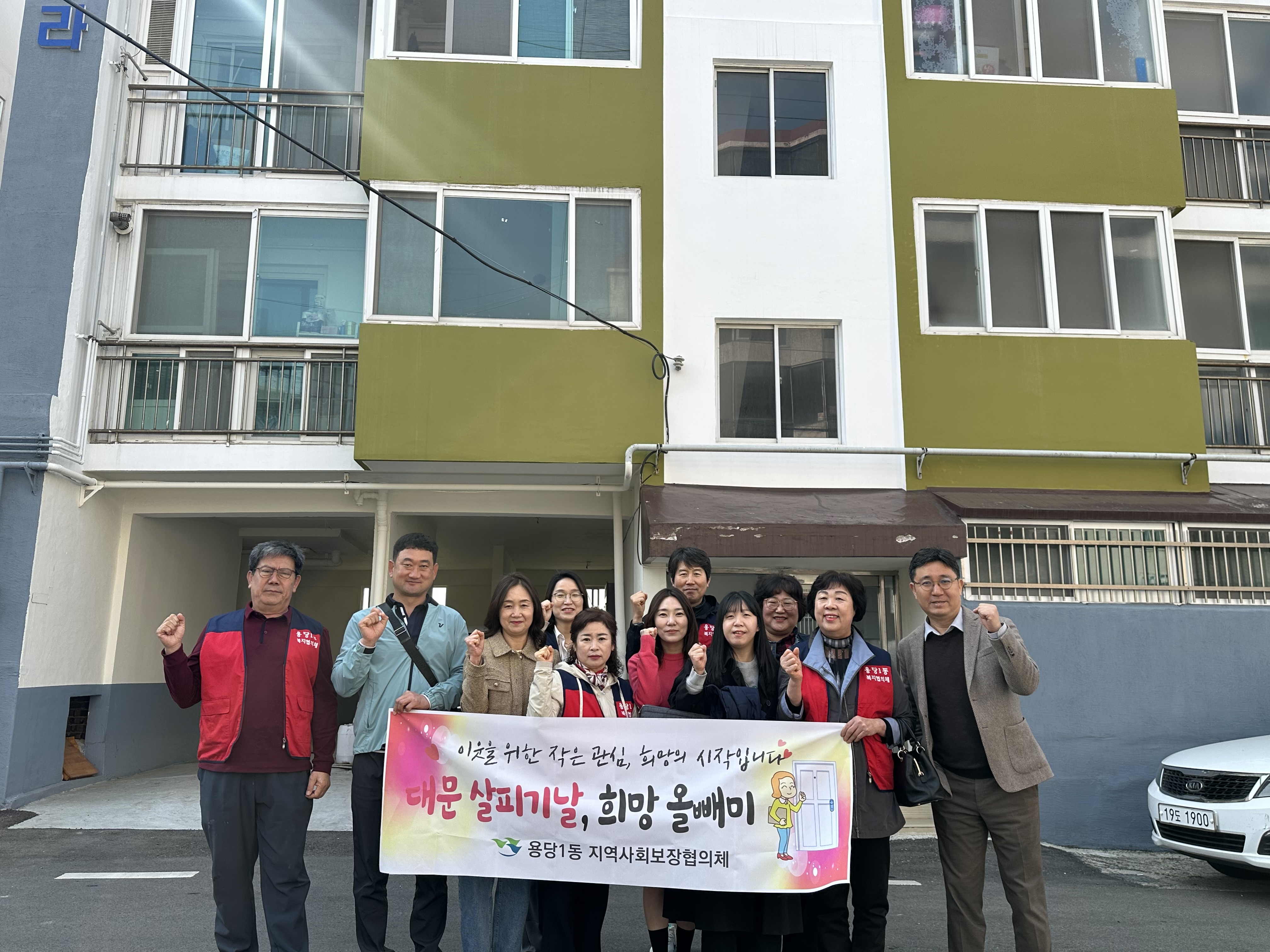 연두색과 흰색이 조화를 이루는 아파트 앞에서, 용당1동 지역사회보장협의체 위원 11명이 '이웃을 위한 작은 관심, 희망의 시작입니다. 대문 살피기날, 희망 울빼미 용당1동 지역사회보장협의체' 문구가 적힌 현수막을 들고 단체 기념촬영을 하고 있는 모습. 참여자들은 주먹을 들어 다짐의 포즈를 취하고 있다.