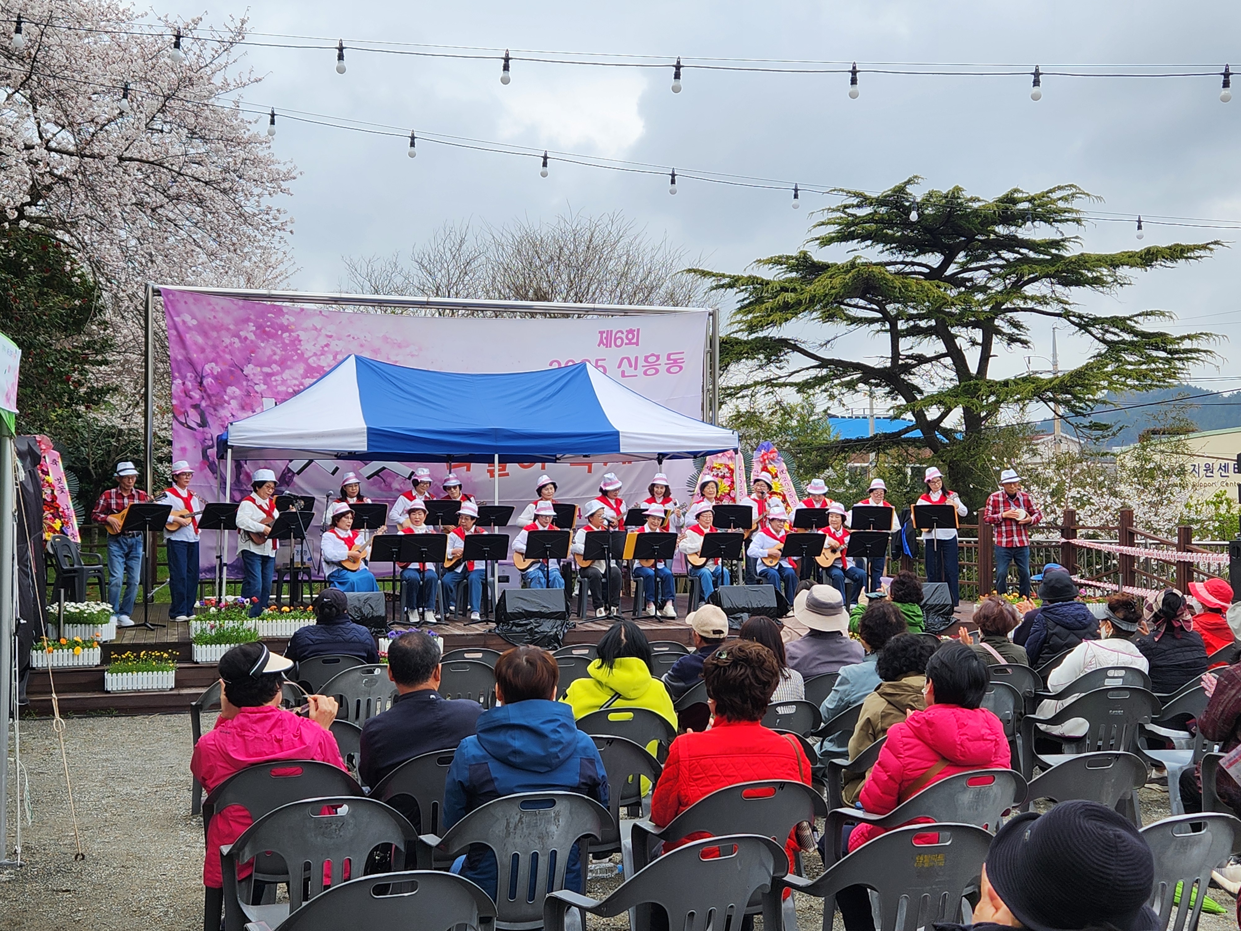‘제6회 2025 신흥동 벚꽃 나들이 축제’ 행사에서 시민 공연팀이 무대 위에서 악기를 연주하고 있는 모습. 단체복을 맞춰 입은 연주자들이 줄지어 서 있으며, 관람객들은 야외 의자에 앉아 무대를 감상 중이다.