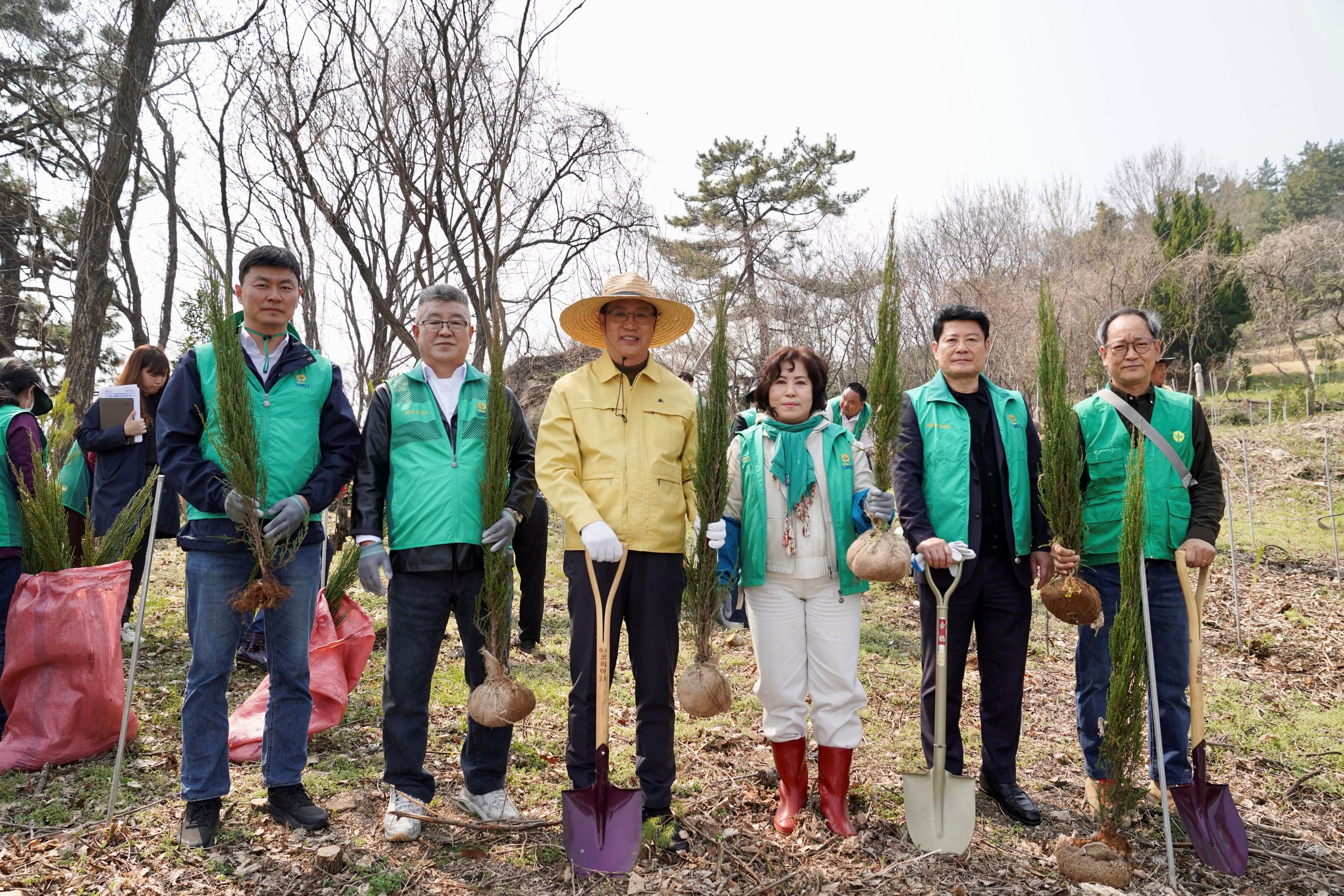 야외에서 여러 사람이 일렬로 서서 삽과 묘목을 들고 기념 촬영을 하고 있는 모습이다. 뒤쪽에는 나무와 흙밭이 보인다.