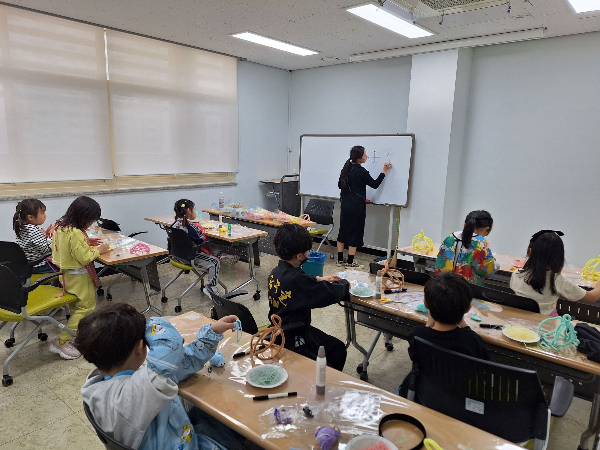 실내 교실에서 여러 어린이가 책상에 앉아 활동을 하고 있는 모습이다. 앞쪽에는 한 사람이 화이트보드에 그림을 그리며 설명하고 있다. 책상 위에는 다양한 만들기 재료와 도구가 놓여 있으며, 어린이들이 손으로 재료를 만지며 작업을 하고 있다.