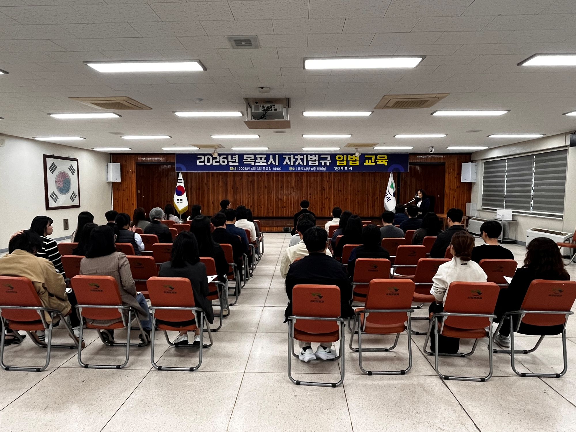 실내 강의실에서 여러 사람이 의자에 앉아 교육을 듣고 있는 모습이다. 앞쪽에는 발표자가 서서 강의를 진행하고 있으며, 뒤쪽에는 ‘2026년 목포시 자치법규 입법 교육’이라고 적힌 현수막이 걸려 있다. 강의실에는 태극기와 기관 깃발이 세워져 있다.