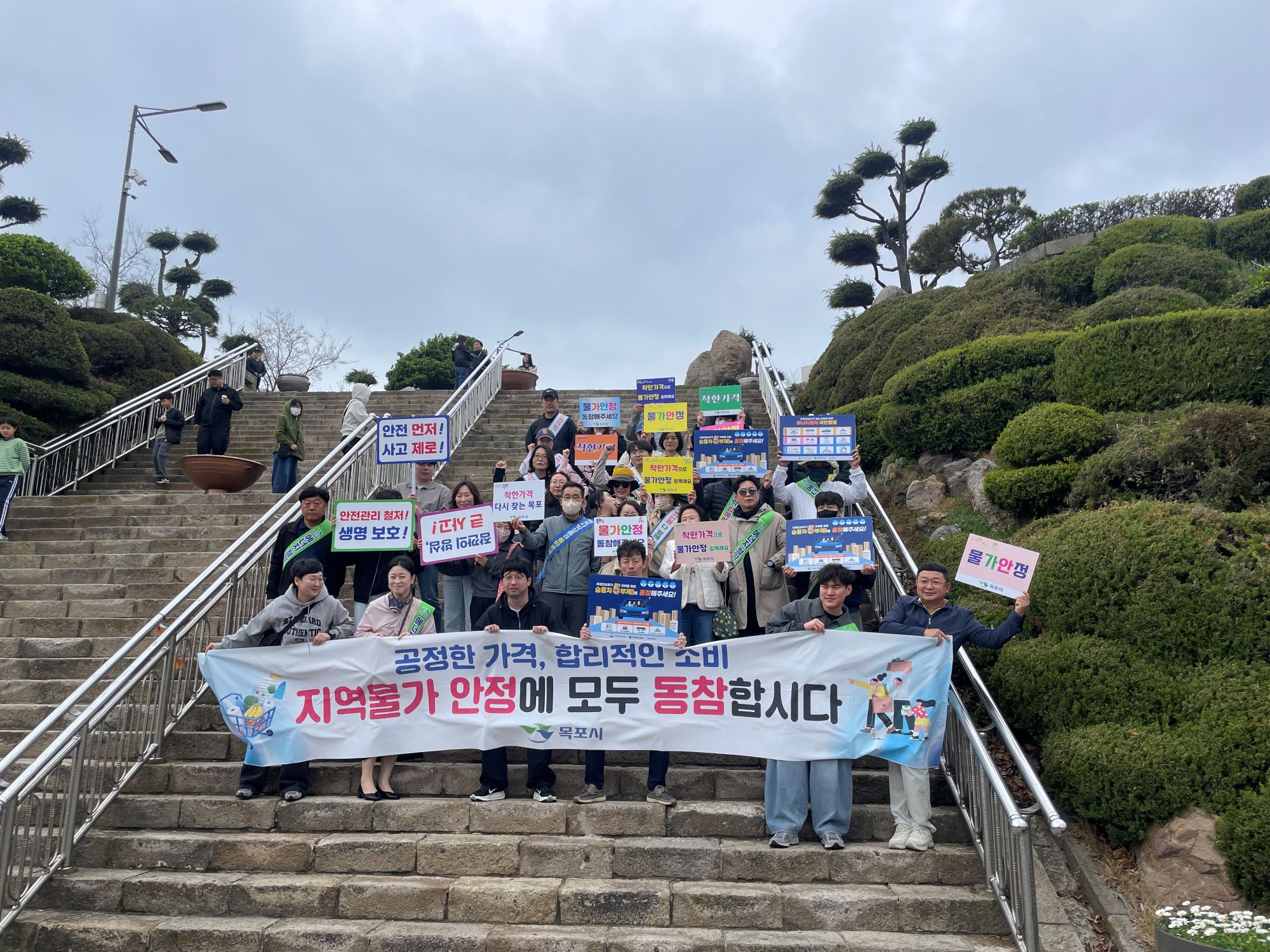 야외 계단에서 여러 사람이 모여 기념 촬영을 하고 있는 모습이다. 앞쪽에는 ‘공정한 가격, 합리적인 소비 지역물가 안정에 모두 동참합시다’라고 적힌 현수막이 보인다. 사람들이 들고 있는 피켓에는 ‘안전 먼저 사고 제로!’, ‘안전관리 철저! 생명 보호!’, ‘착한가격 업소 이용하기’, ‘물가안정 동참해 주세요’, ‘물가안정’, ‘착한가격 다시 찾은 행복’ 등의 문구가 보인다. 주변에는 계단과 난간, 나무와 조경이 있는 야외 공간이 보인다.
