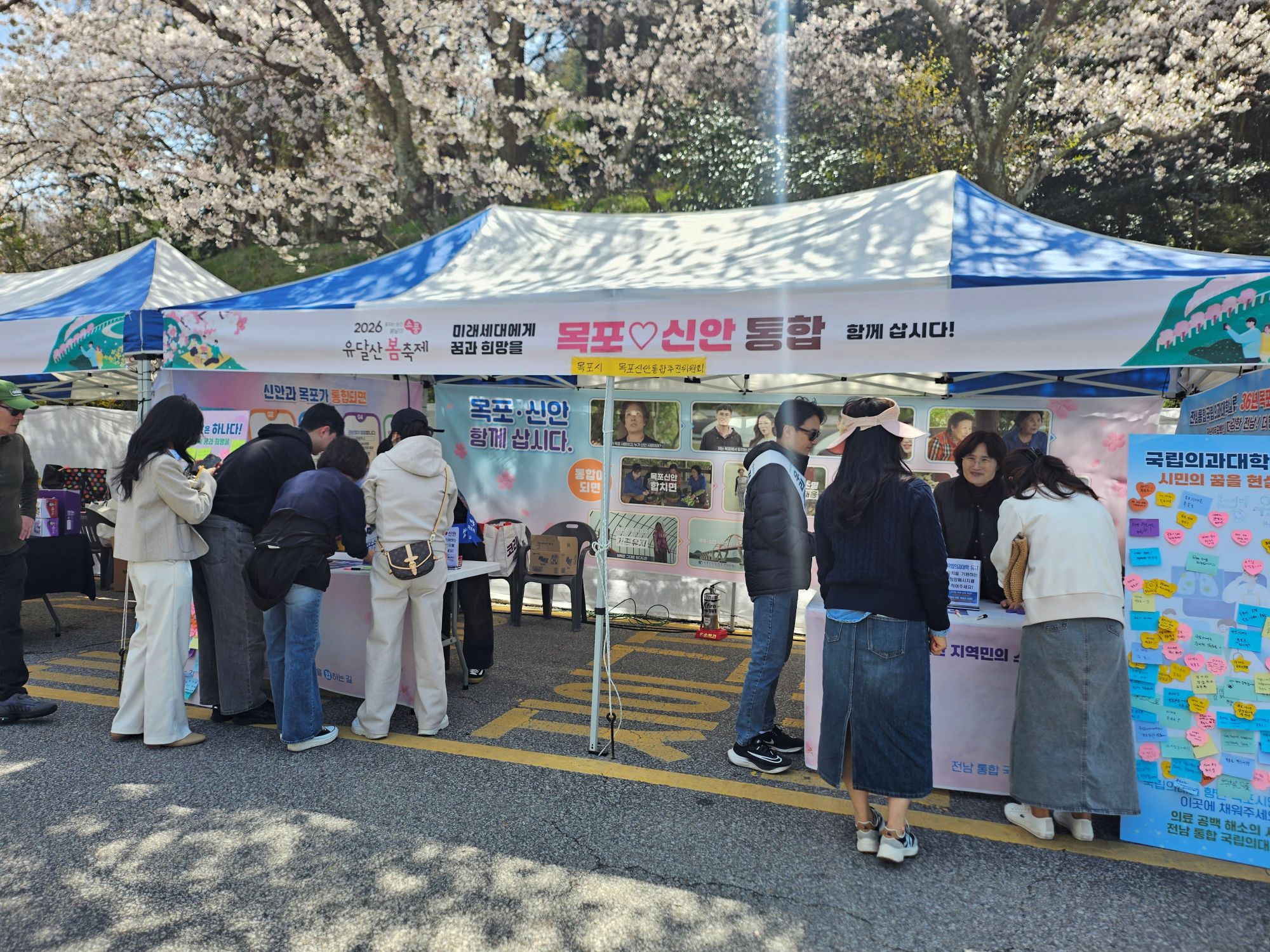 야외 행사 부스에서 여러 사람이 참여하고 있는 모습이다. 천막에는 ‘2026 유달산 봄축제’, ‘미래세대에게 꿈과 희망을’, ‘목포♡신안 통합 함께 삽시다!’라는 문구가 적혀 있다. 부스 내부에는 ‘목포·신안 함께 삽시다.’라는 안내판과 다양한 홍보물이 전시되어 있으며, 한쪽에는 ‘국립의과대학 30년의 꿈을 현실로’라는 문구가 적힌 게시판이 보인다. 여러 사람이 테이블 앞에서 서서 안내를 보거나 참여 활동을 하고 있으며, 게시판에는 여러 개의 메모지가 붙어 있다.