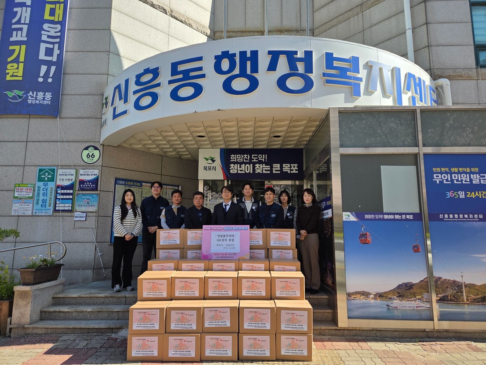 건물 출입구 앞에서 여러 사람이 서서 기념 촬영을 하고 있는 모습이다. 건물 상단에는 ‘신흥동 행정복지센터’라고 적힌 간판이 보이며, 출입구 안쪽에는 ‘희망찬 도약! 청년이 찾는 큰 목포’라는 문구가 보인다. 앞쪽에는 여러 개의 상자가 쌓여 있으며, 중앙 안내판에는 ‘성금품 전달식’과 ‘60상자’라는 문구가 표시되어 있다. 상자마다 동일한 안내 문구가 부착되어 있다. 건물 한쪽에는 ‘무인 민원 발급 365일 24시간’이라는 안내 문구가 보인다.