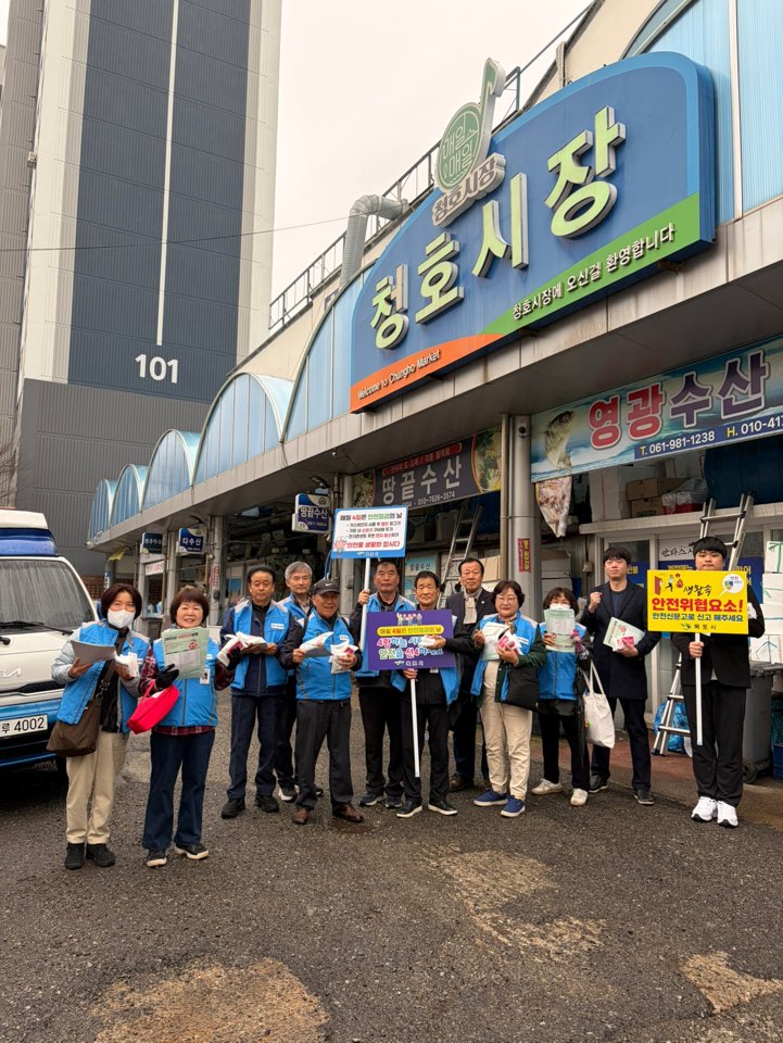 야외 시장 입구에서 여러 사람이 서서 안내 활동을 하고 있는 모습이다. 뒤쪽 건물에는 ‘청호시장’과 ‘청호시장 오신걸 환영합니다’라고 적힌 간판이 보인다. 사람들이 들고 있는 안내물에는 ‘안전위험요소! 안전신문고로 신고하세요’라는 문구가 보이며, 일부는 전단지를 들고 있다. 주변에는 상가 간판과 차량이 보인다.