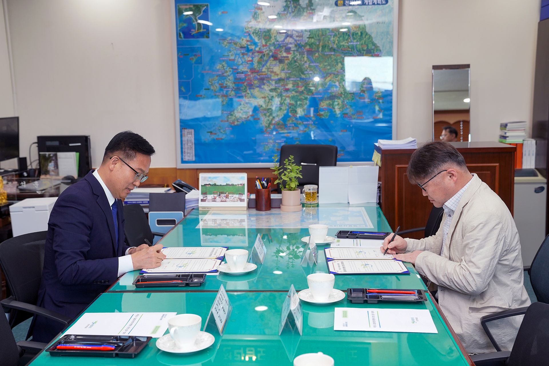 실내 회의 공간에서 두 사람이 테이블에 마주 앉아 문서에 서명을 하고 있는 모습이다. 테이블 위에는 서류와 펜, 컵 등이 놓여 있으며, 뒤쪽 벽에는 지도와 사무용 물품이 보인다.