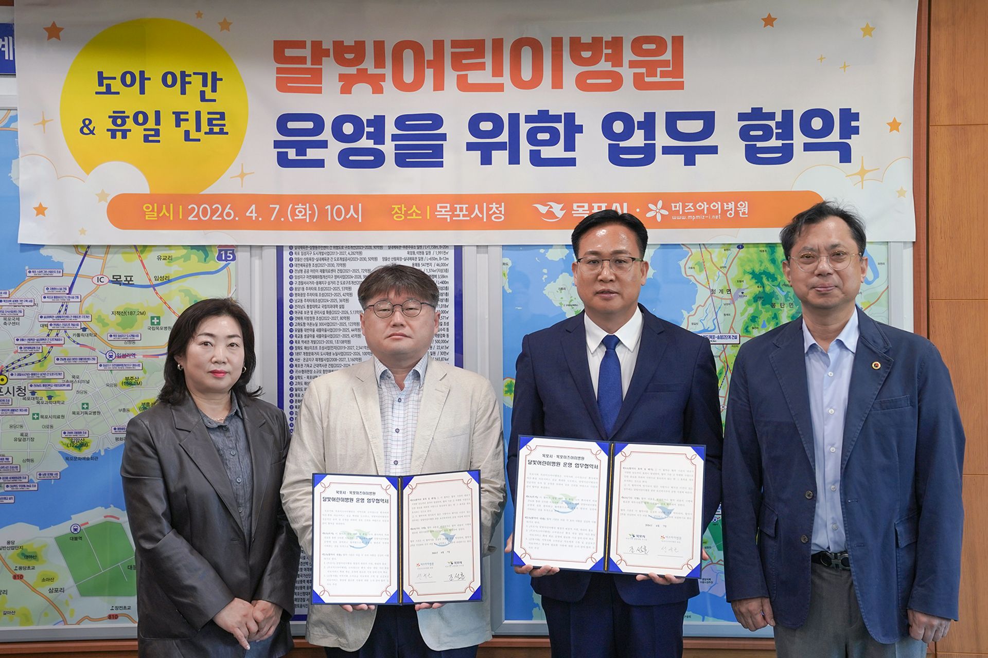 실내에서 여러 사람이 나란히 서서 문서를 들고 기념 촬영을 하고 있는 모습이다. 뒤쪽에는 ‘달빛어린이병원 운영을 위한 업무 협약’이라고 적힌 현수막이 걸려 있으며, 왼쪽에는 ‘소아 야간 & 휴일 진료’라는 문구가 보인다. 현수막에는 ‘일시 2026. 4. 7.(화) 10시’, ‘장소 목포시청’, ‘목포시 · 미즈아이병원’이라는 내용이 표시되어 있다.