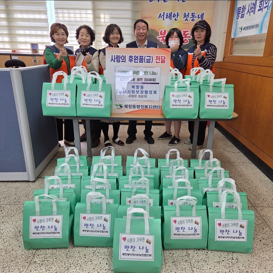 실내 공간에서 여러 사람이 테이블 뒤에 서서 기념 촬영을 하고 있는 모습이다. 테이블 위와 바닥에는 여러 개의 가방이 놓여 있으며, 가방에는 ‘사랑이 가득한 반찬 나눔’이라고 적힌 문구가 보인다. 중앙에는 ‘사랑의 후원품(금) 전달’이라고 적힌 안내판이 있으며, 그 아래에 ‘「사랑이 듬뿍! 반찬 나눔」 밑반찬 3종 세트 (50세대)’와 ‘북항동 지역사회보장협의체 위원장 최상식’, ‘북항동행정복지센터’라는 문구가 표시되어 있다. 뒤쪽 현수막에는 ‘서해안 첫동네 북항동’이라는 문구가 보인다.