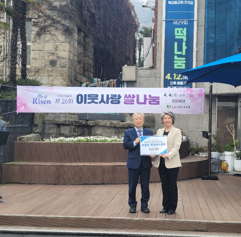 야외 무대에서 두 사람이 서서 기념 촬영을 하고 있는 모습이다. 뒤쪽에는 ‘He is Risen 2026 부활절 제26회 이웃사랑 쌀 나눔’이라고 적힌 현수막이 걸려 있다. 현수막 오른쪽에는 ‘4. 4.(토) 오후 2:00, 500세대’라는 문구가 보인다. 두 사람은 ‘유달동 행정복지센터 (55가구)’라고 적힌 안내판을 함께 들고 있다. 주변에는 건물과 안내 표지판이 보인다.