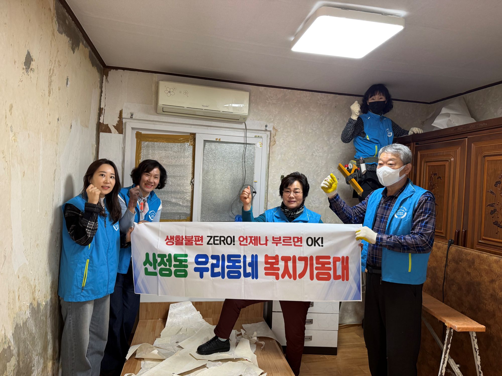 실내 공간에서 여러 사람이 함께 벽지 작업을 진행하고 있는 모습이다. 사람들이 파란색 조끼를 입고 있으며, 한 사람이 벽지 제거 작업을 하고 있고 다른 사람들은 손을 들어 보이며 서 있다. 앞쪽에는 ‘생활불편 ZERO! 언제나 부르면 OK! 산정동 우리동네 복지기동대’라고 적힌 현수막을 들고 있다. 바닥에는 제거된 벽지 조각이 놓여 있고, 실내에는 가구와 생활용품이 보인다.