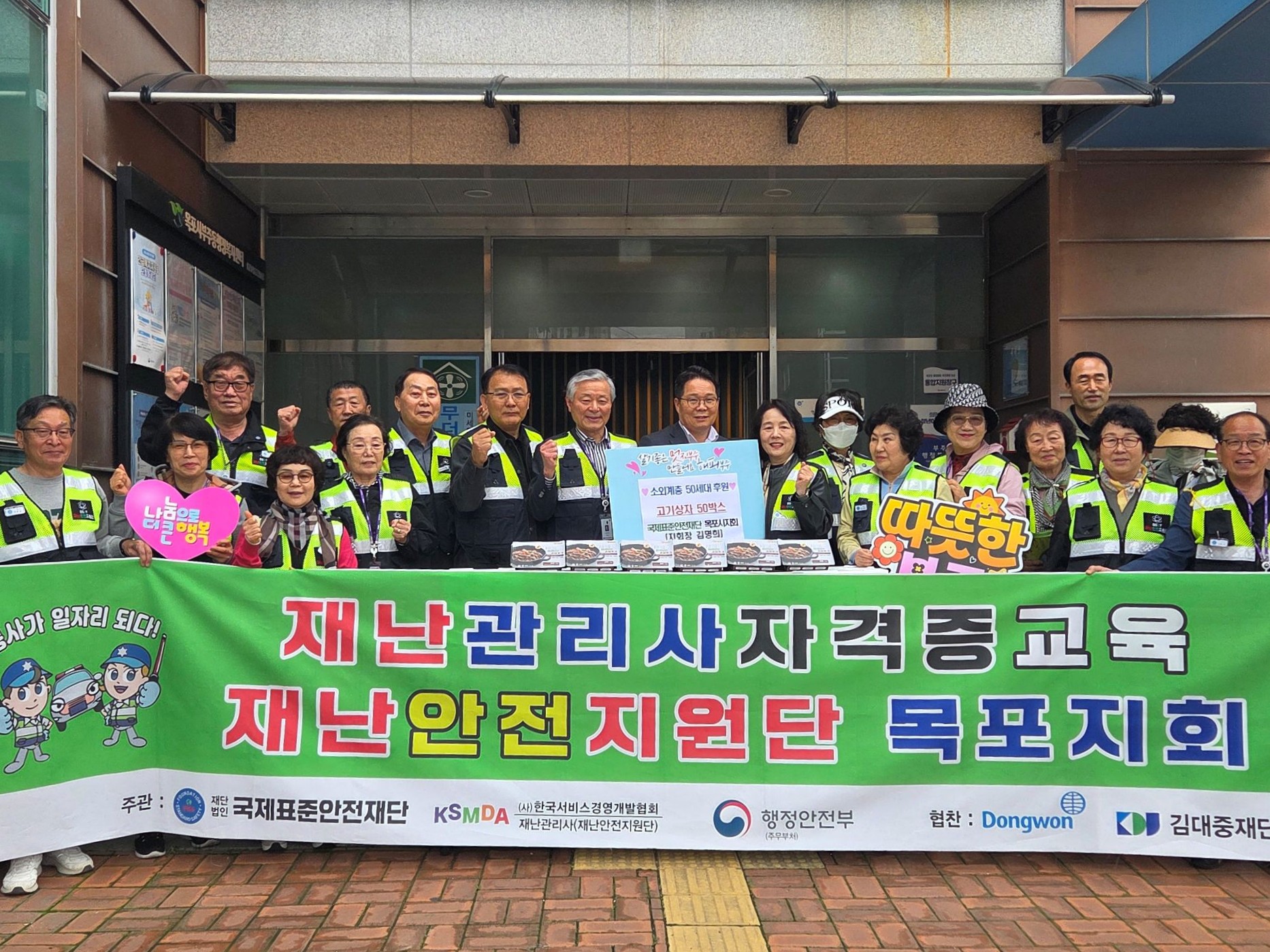 재난관리사자격증교육 재난안전지원단 목포지회 문구가 적힌 현수막 뒤로 소외계층에 후원하기 위한 고기상자가 있다. 주변으로 화이팅을 하며 웃으며 카메라를 바라보고 있는 19명의 단체사진