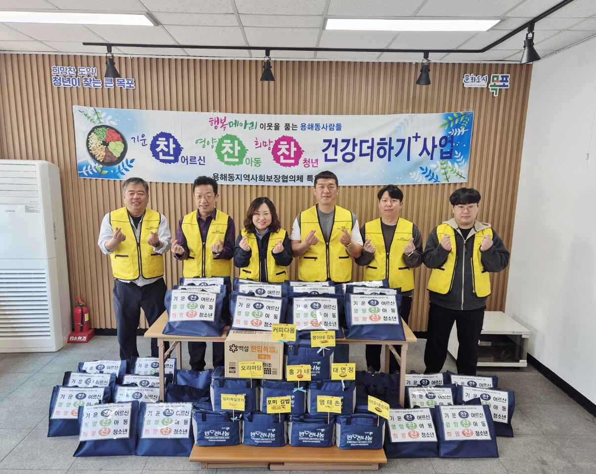 희만찬 도약! 청년이 찾는 큰 목포 문화도시 목포. 행복메아리 이웃을 품는 용해동사람들. 기운찬 어르신, 영양찬 아동, 희망찬 청년 건강더하기 사업 용해동지역사회보장협의체 현수막이 걸려있다. 후원 물품이 앞에 나열되어 있고, 그 뒤로 양손으로 손하트를 한 6명의 직원들이 있다