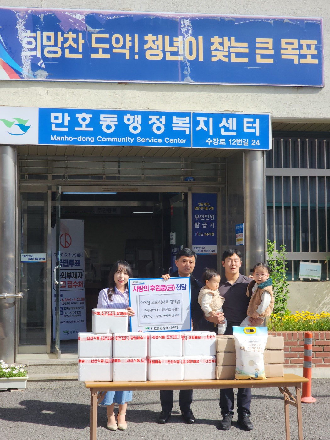 희망찬 도약@ 청년이 찾는 큰 목포 만호동행정복지센터 앞에서 사랑의 후원품(금)전달 기념 사진. 스티로폼에 담긴 김치 9박스와 쌀포대 뒤로 6개의 박스가 놓여있다. 여성과 중년 남성이 전달 물품이 적힌 안내팻말을 들고 있으며, 젛은 남성이 미취학 아동 두명을 품에 안고 있다