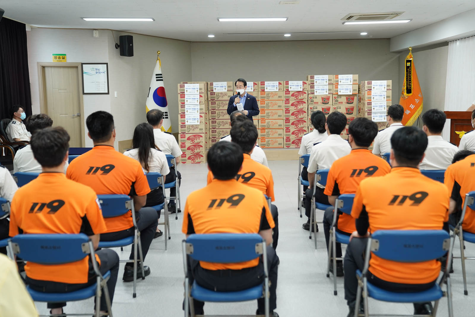 소방대원들이 앉아 있고 앞에 서서 시장님이 연설하고 있다 뒤로 기념품이 보인다