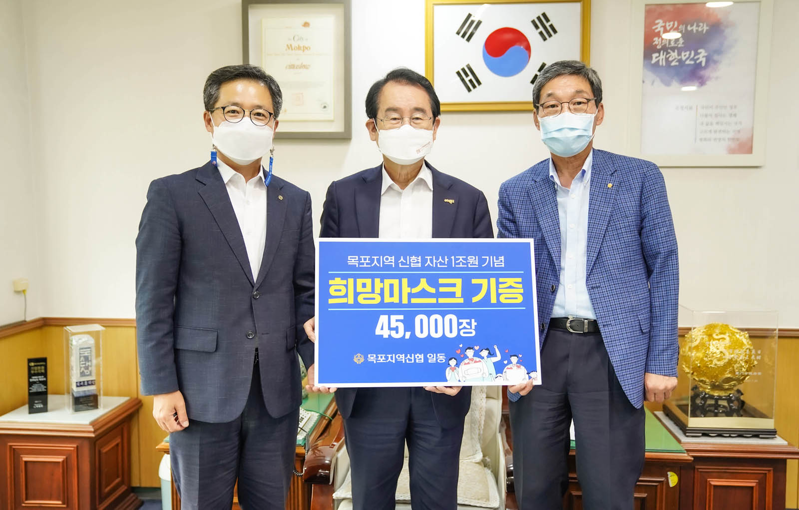 목포지역 신협 자산 1조원 기념 희망마스크 기증 45,000장 목포지역신협 일동 이라고 적힌 배너를 들고 시장님과 관계자 두 명이 기념사진을 찍고 있다.