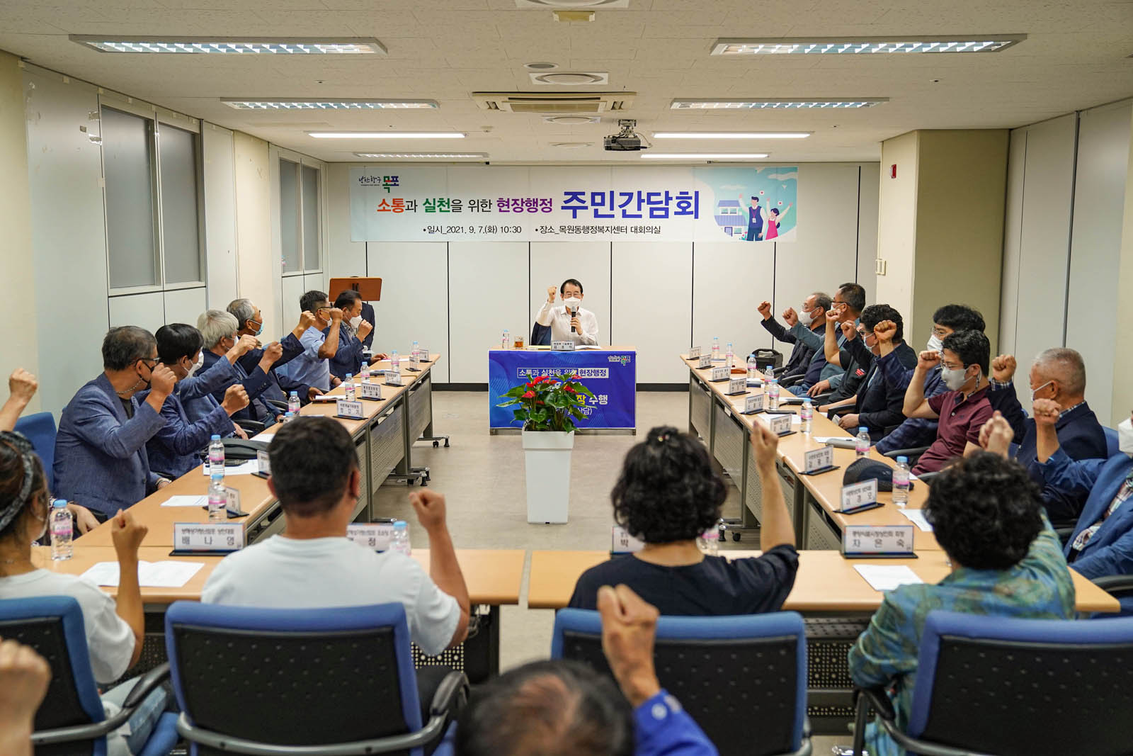 소통과 실천을 위한 현장행정 주민간담회 라고 적힌 현수막이 걸려 있고 시장님이 앞에 앉아 마이크를 들고 관계자들과 함께 화이팅하고 있는 사진이다.