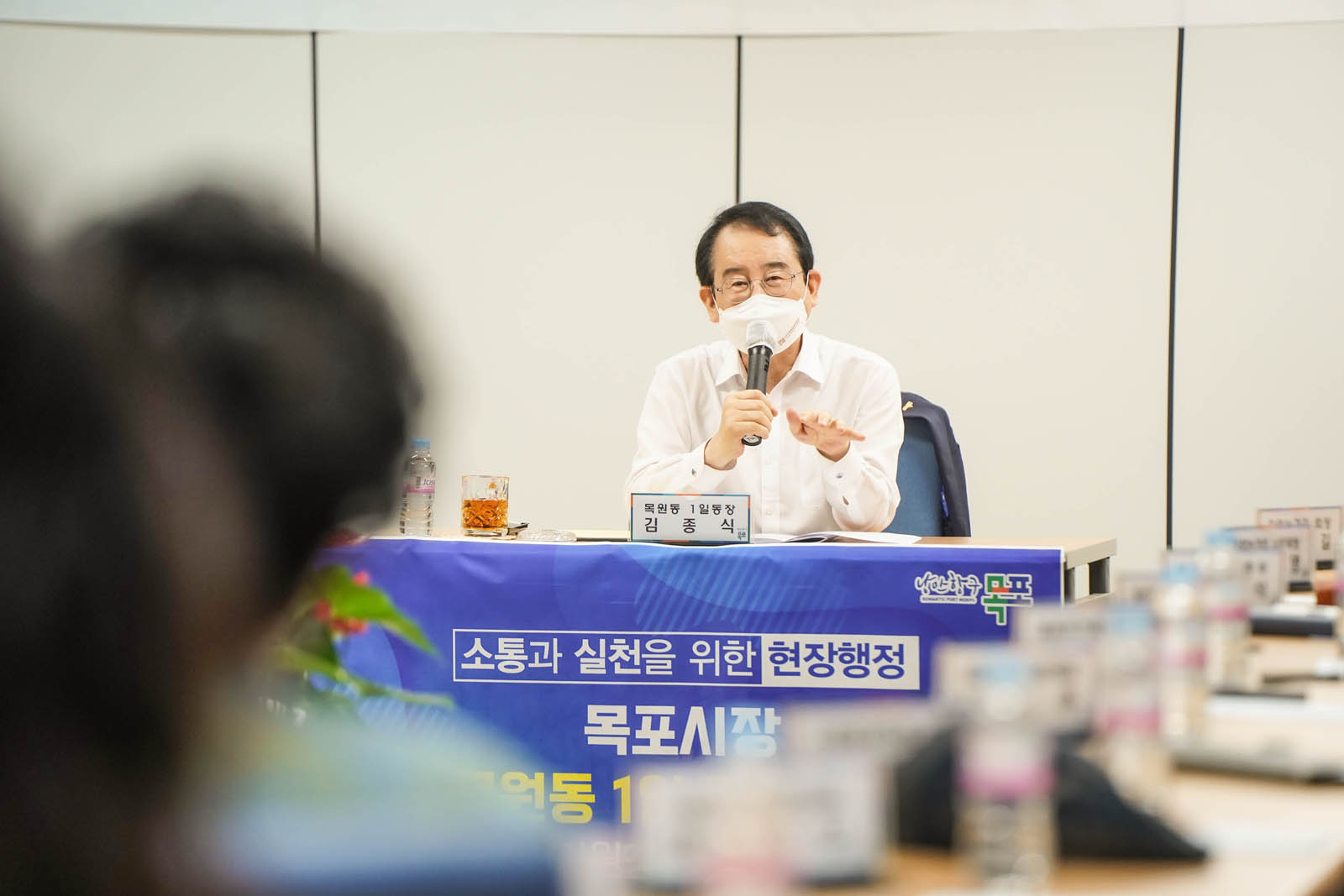 시장님이 마이크를 들고 이야기하고 있다. 탁자에는 목원동 1일동장 김종식이라고 적힌 작은 명패가 놓여 있다.