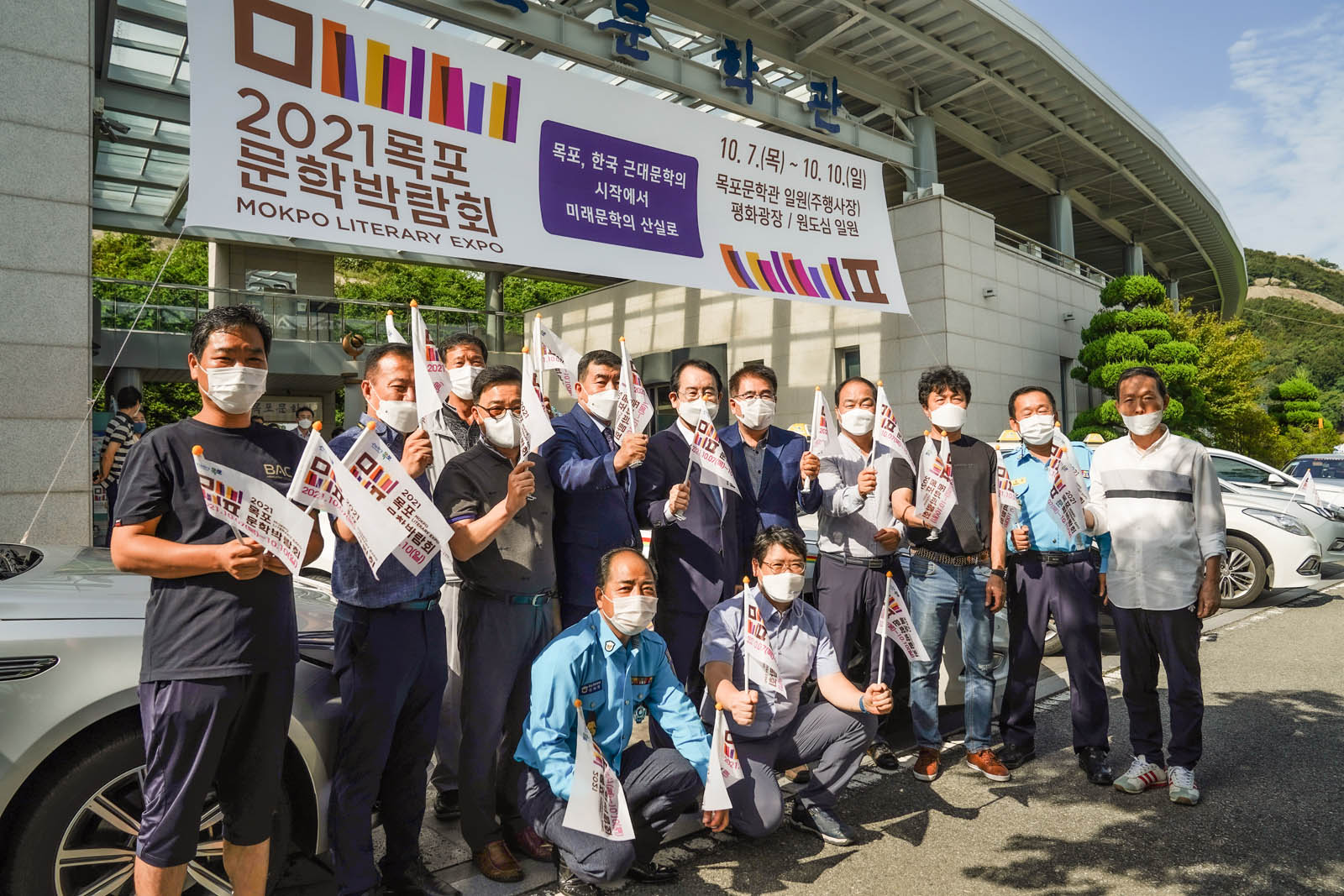 2021목포문학박람회 목포, 한국 근대문학의 시작에서 미래 산실로 10.7(목)~10.10(일) 목포문학관 일원(주행사장) 평화광장/원도심 일원 이라고 적힌 현수막이 목포문학관 앞에 걸려 있고 그 앞으로 여러사람들이 홍보깃발을 들고 기념촬영중이다.