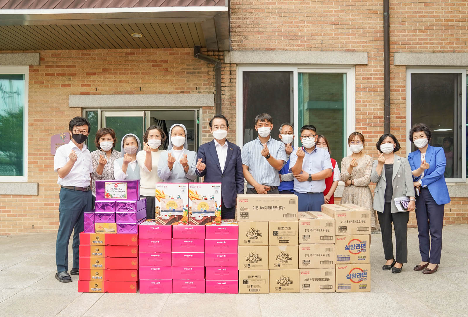 수녀님과 복지시설 관계들 여렷이 기념품을 앞에 놓고 손가락하트를 하며 기념촬영을 하고 있다