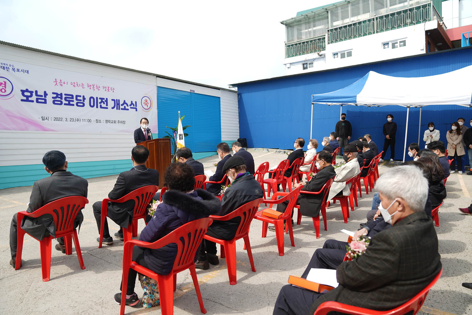(03.23.연락교회주차장) 목원동 호남경로당 이전 개소식에서 목포시장이 연설중이고 참석자들이 의자에 착석해서 경청하고 있다.