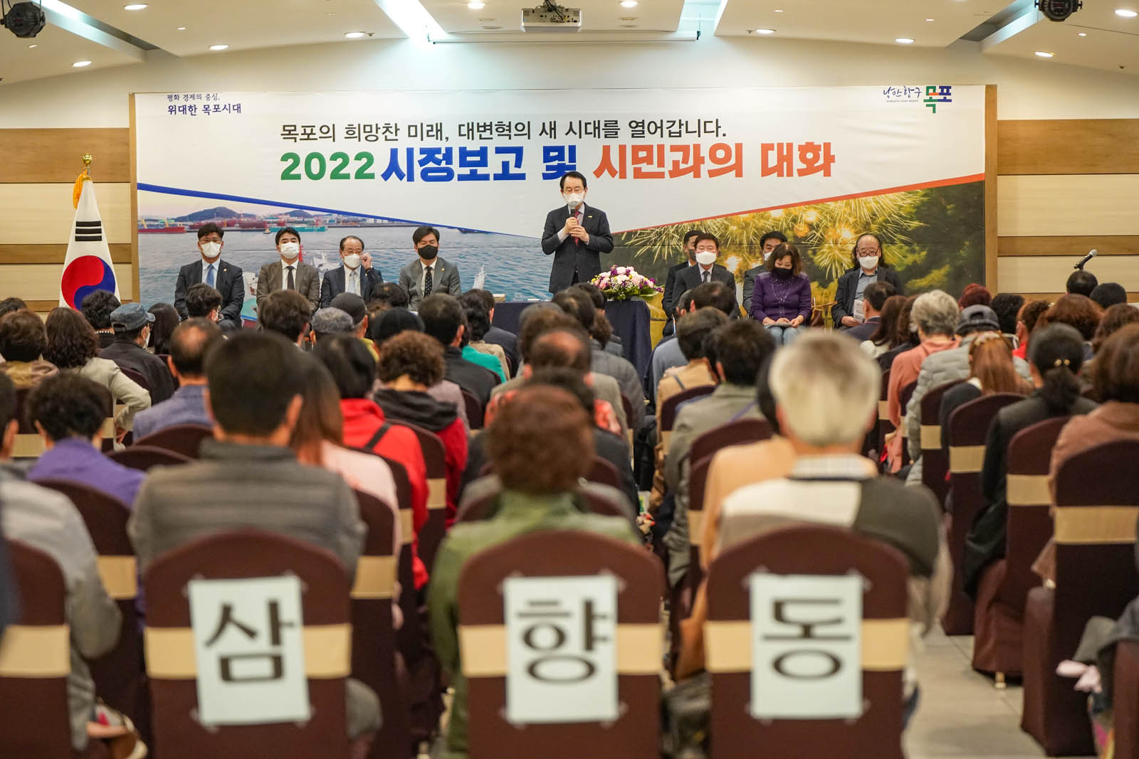 (03.30.에메랄드웨딩홀) 시민들이 자리에 착석해 있고 시장이 앞에 서서 연설 중인 사진이다.