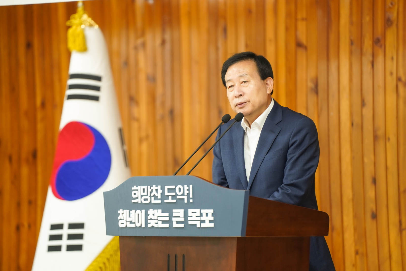 (07.04.회의실) 직원 정례조회를 진행하는 모습을 뒤에서 찍은 사진이다. 시장이 단상에서 연설 중이고 참석자들은 의자에 착석해서 경청하고 있다.