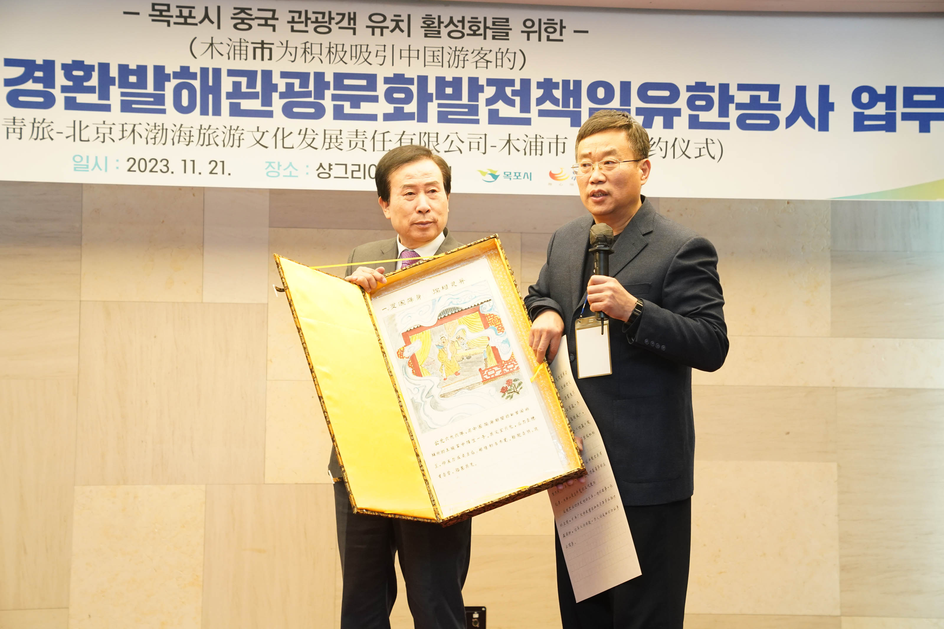 목포시 중국 관광객 유치 활성화를 위한 목포시 중청여 북경환발해관광문화발전책임유한공사 업무 협약식 플랜카드를 뒤에두고 단상위에서 박홍률 목포시장이 금색상자를 들고있고 사회자가 설명하는 모습