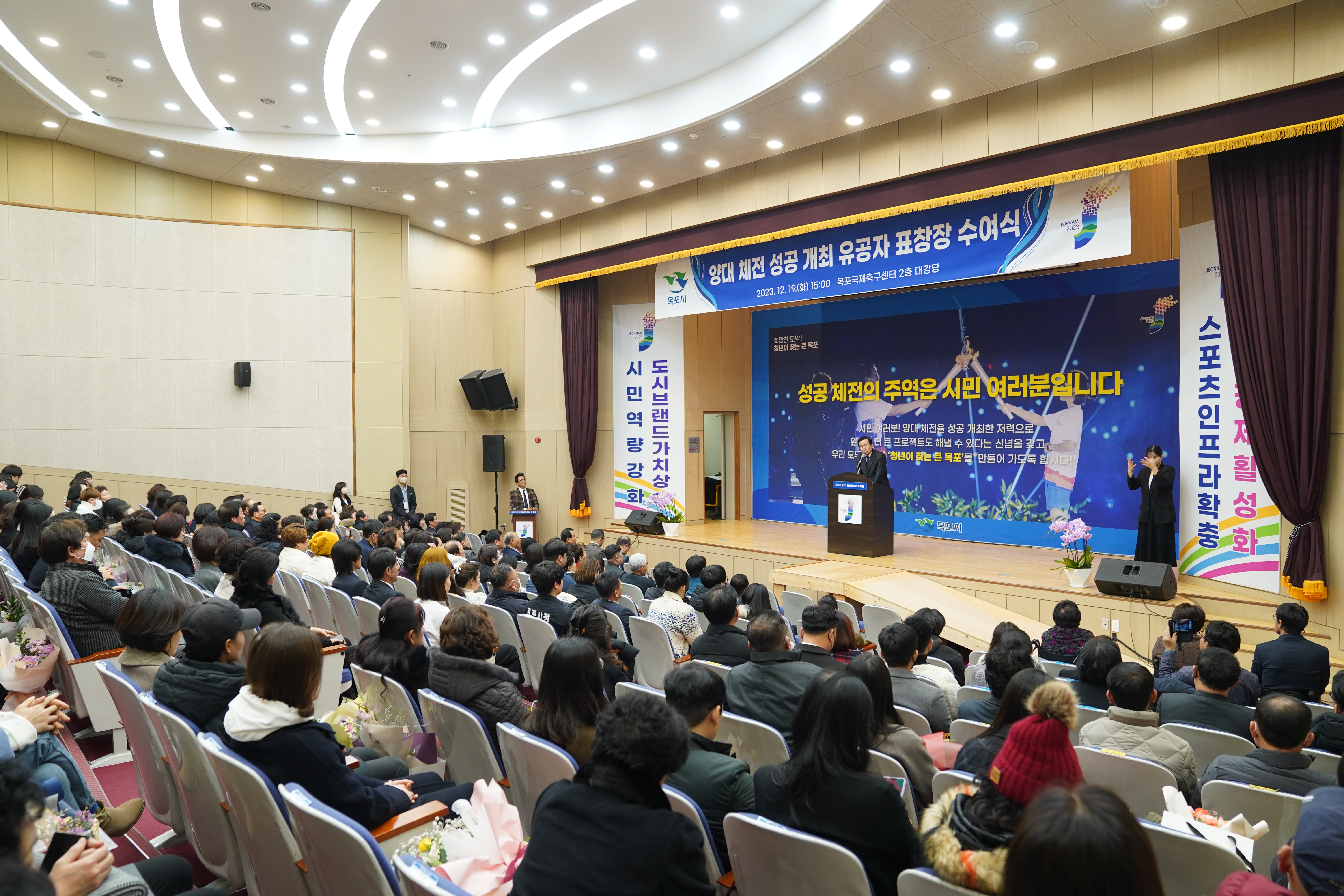 성공 체전의 주역은 시민여러분입니다. 글이 적힌 배경을 두고 양대 체전 성공 개최 유공자 포창장 수여식 플랜카드가 위에 걸려있고 연설하는 목포시장 박홍률과 경청하는 청중