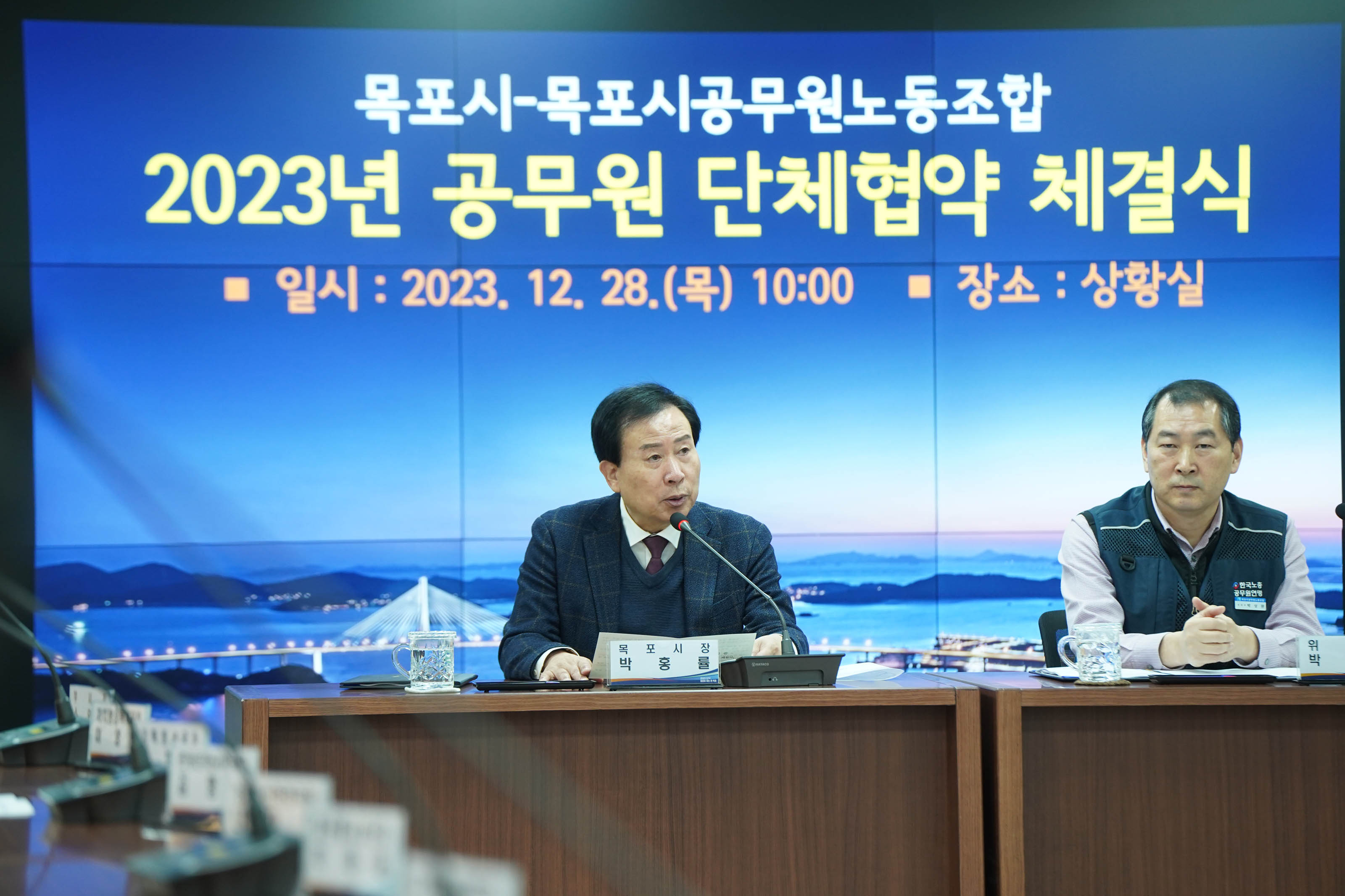 목포시 목포시공무원노동조합 2023 공무원 단체협약 체결식 일시 : 2023.12.28(목) 10:00 장소:상황실이 적혀있는 배경 앞에서 연설중인 목포시장 박홍률과 경청하는 위원장