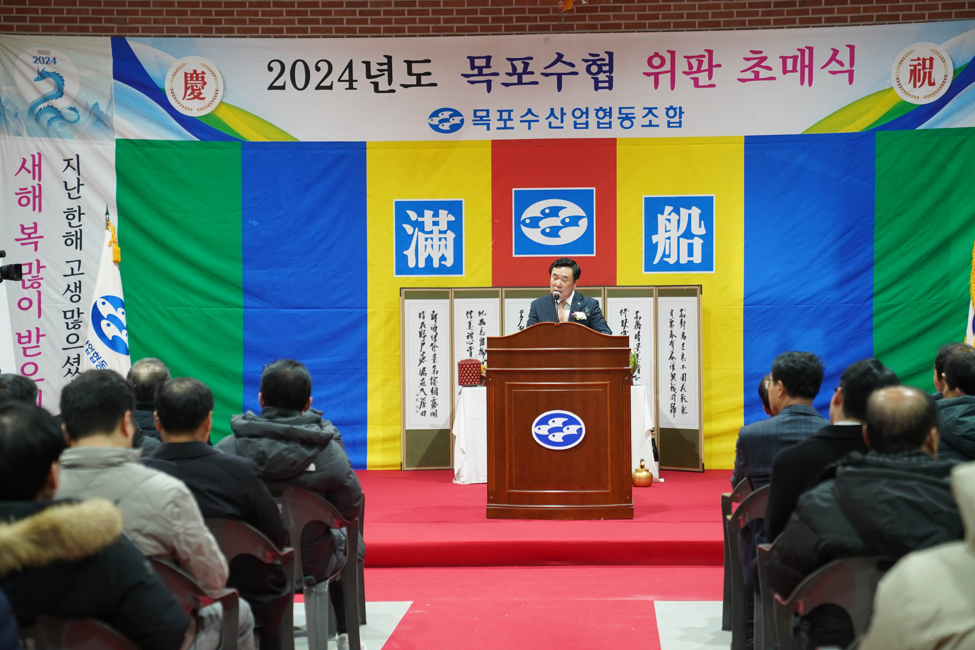 2024년도 목포수협 위판 초매식 목포수산업협동조합 현수막과 지난해 고생많으셨습니다 새해복많이 받으세요 현수막을 배경으로 두고 연단에서 연설중인 관계자대표와 경청하는 청중