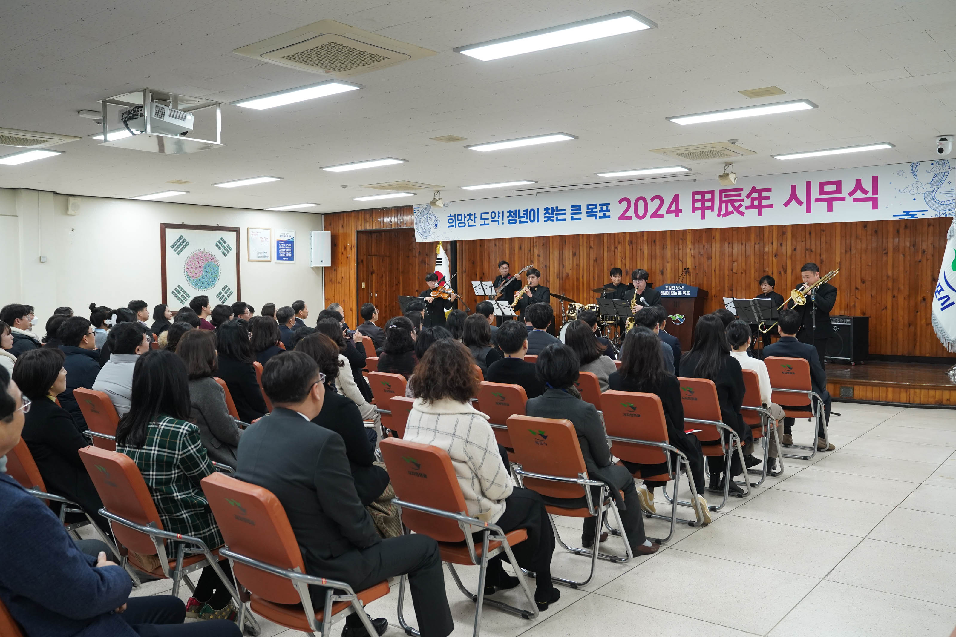 희망찬 도약! 청년이 찾는 큰 목포 2024 甲辰年 시무식 현수막이 걸린 무대위에서 연주중인 사람들과 경청하는 청중들 측면사진