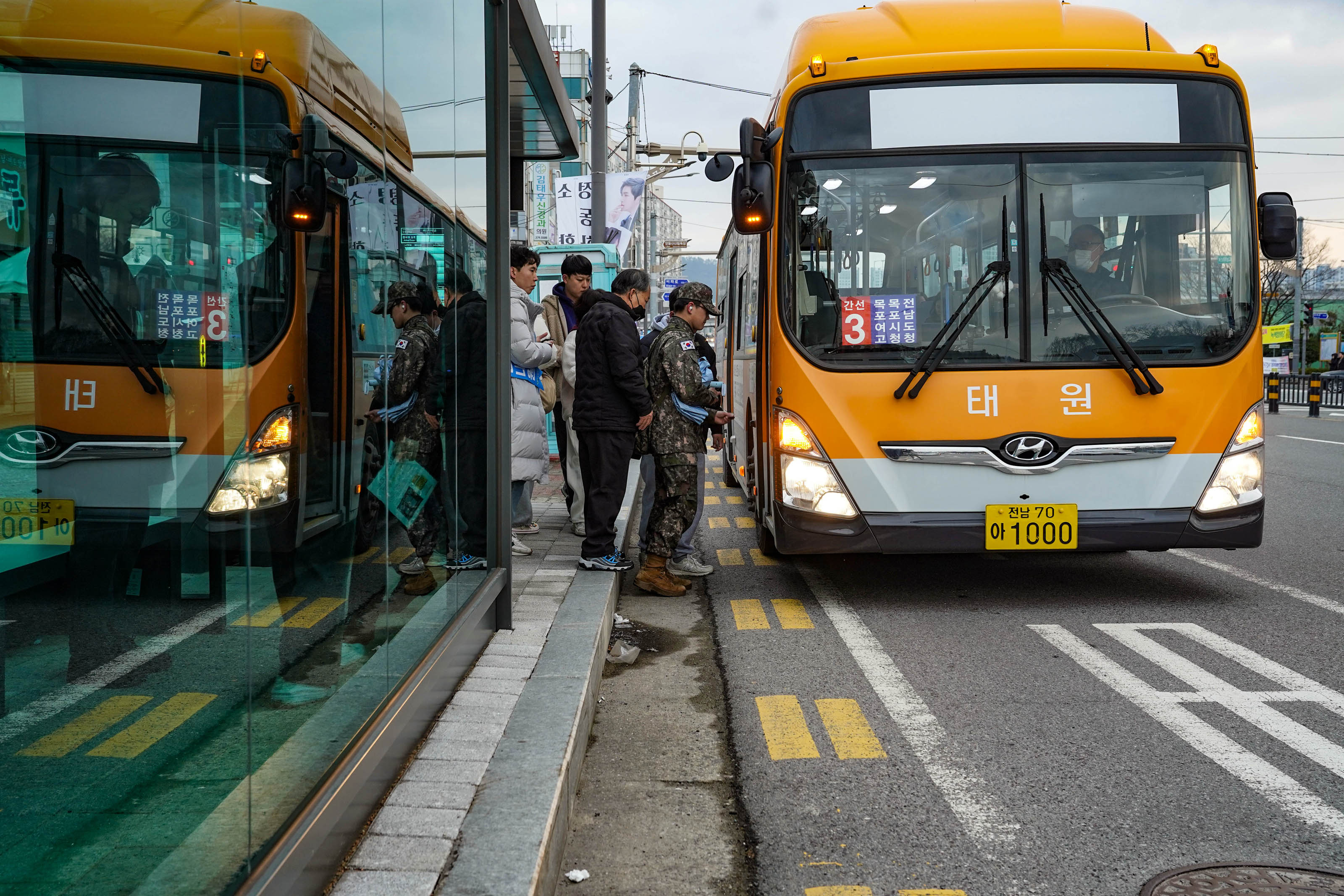 전남 70 아 1000 번호판의 태원 회사의 주황색 버스(간선3, 목포여고, 목포시청, 전남도청)가 정류장 앞에 서있고, 버스를 타기 위해, 군인, 검은색 마스크를 끼고 검은색 의류를 입은 사람들 뒤로 약 3명의 사람들이 줄 서 있다.