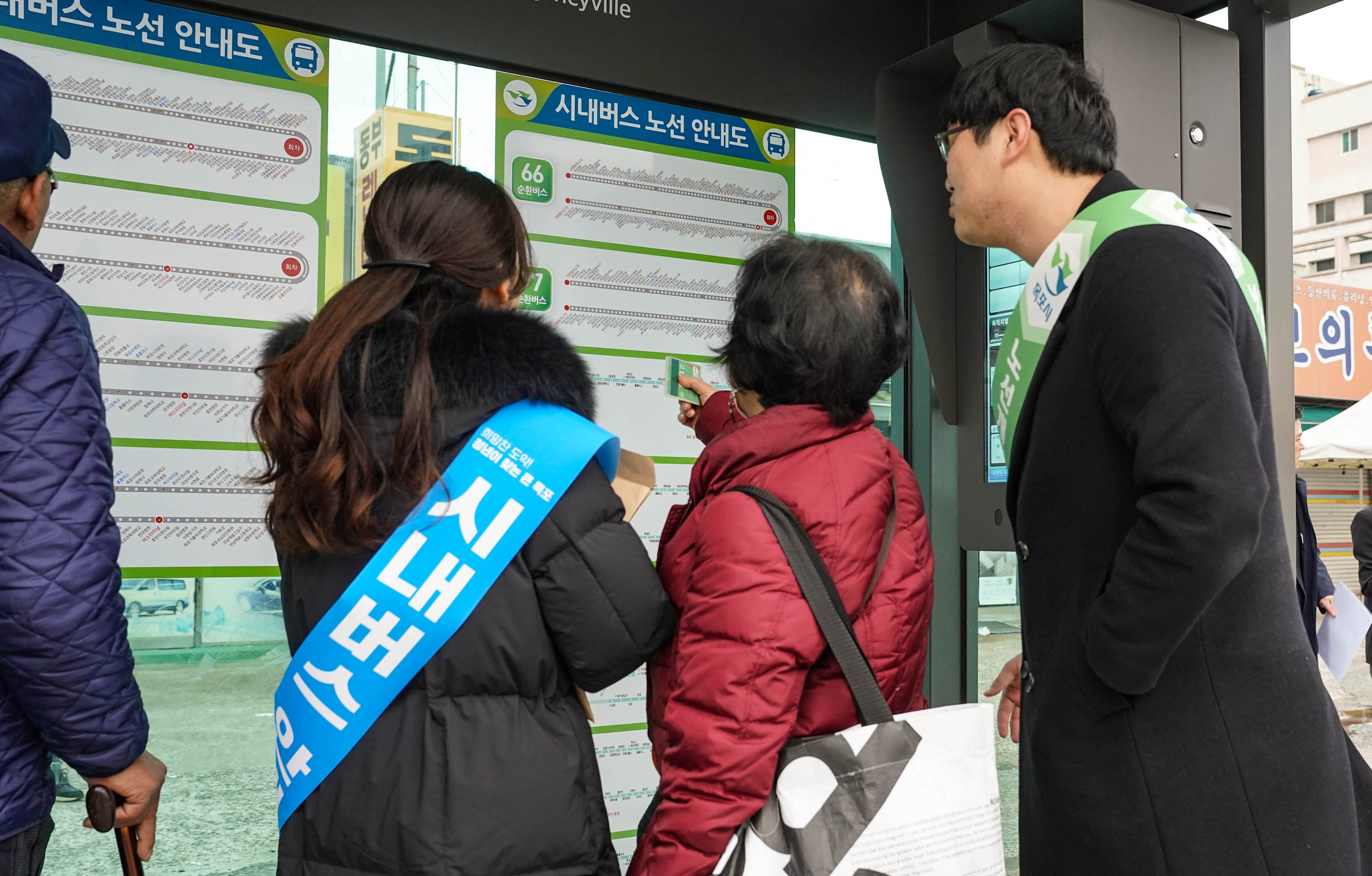 정류장에서 노선을 확인하기 위해 노선 안내도를 보고 있는 자주색 점퍼를 입은 여성과, 파란색 띠(시내버스 안내)를 두른 여성, 연두색 띠(노선개편)를 두른 남성이 양 옆에서 자주색 점퍼를 입은 여성을 도와주는 모습.