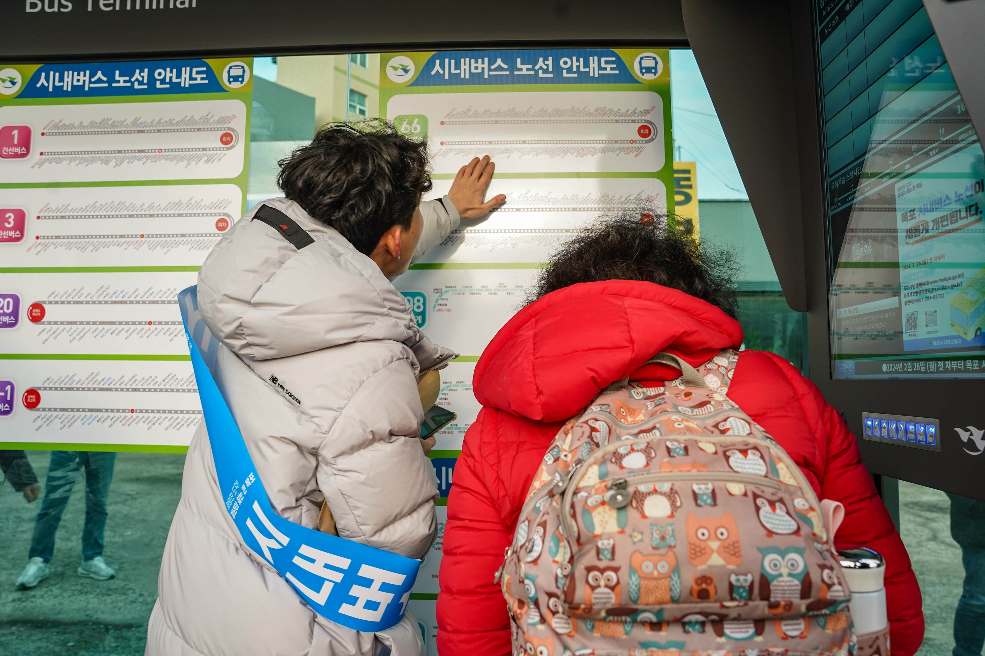 빨간색 점퍼를 입고 부엉이 캐릭터 무늬가 그려진 베이지 색 백팩을 맨 여성이 노선 안내도를 보고 있고, 연회색 패딩을 입고 파란색 띠를 두른 남성이 노선도를 손으로 짚으며 도와주고 있는 모습