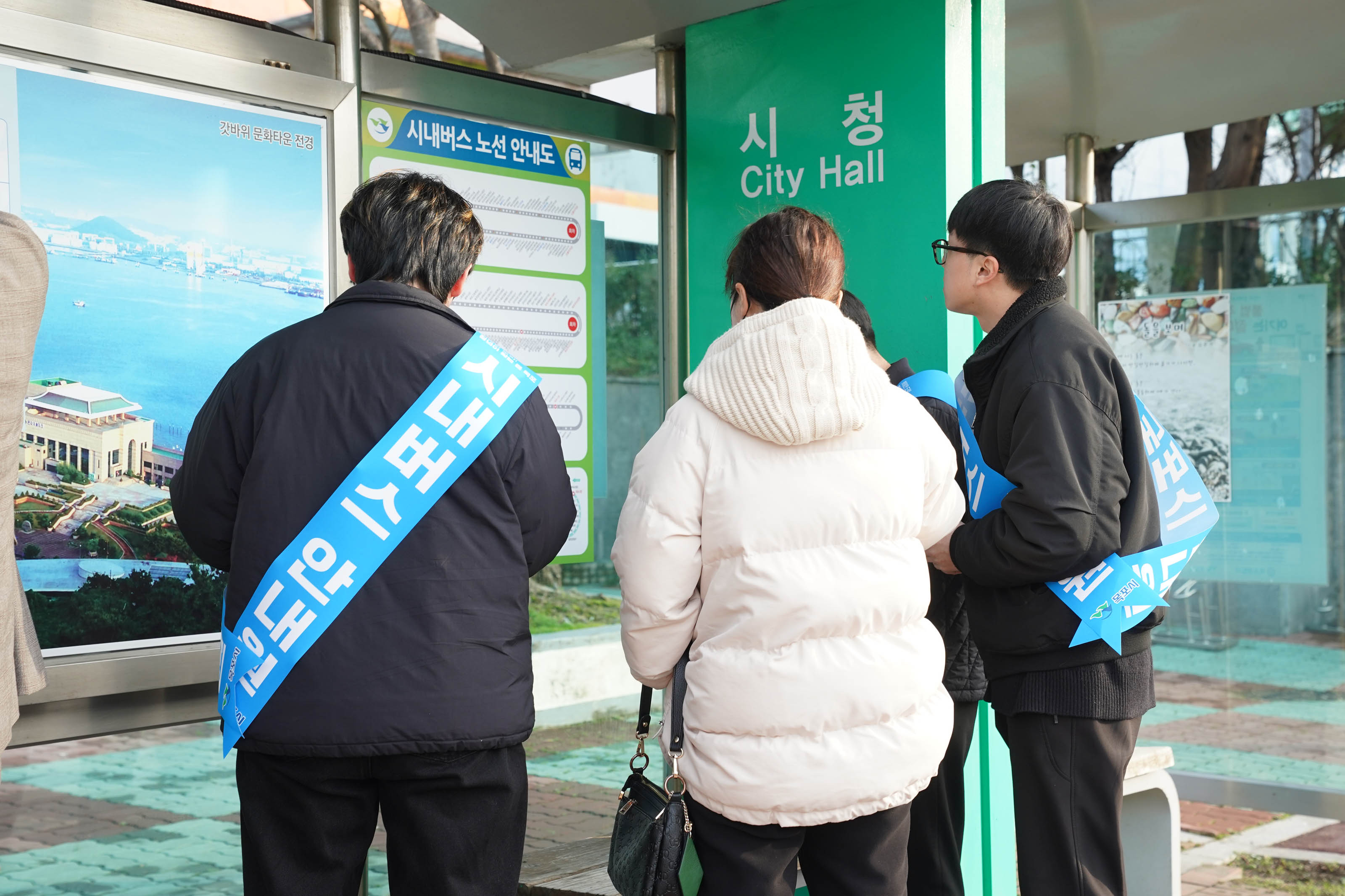시청 Citi Hall 정류 장에서 시내버스 안내도 문구가 적힌 파란색 띠를 두른, 검은색 복장의 남성 3명이 흰색 점퍼를 입고 검은색 미니백을 든 여성을 도와주고 있다