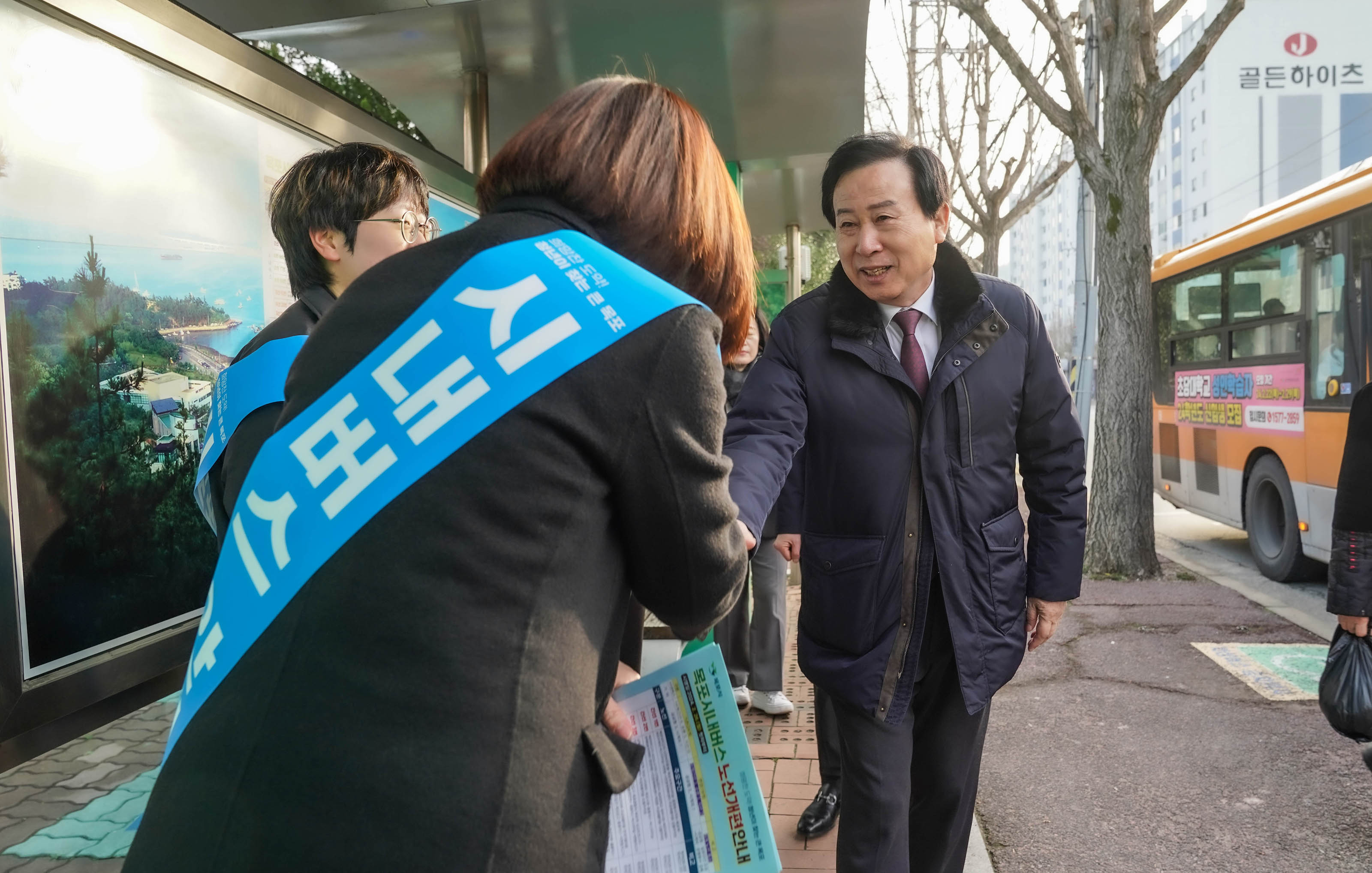 정류장에서 시내버스 안내 문구가 적힌 파란띠를 두른 여성과 악수를 하며 인사하는 박홍률 시장. 여성의 오른손은 악수를 하고 있고, 왼손에는 목포시내버스 노선개편안내 자료를 들고 있다. 도로에는 주황색 버스가 지나가고 있다.