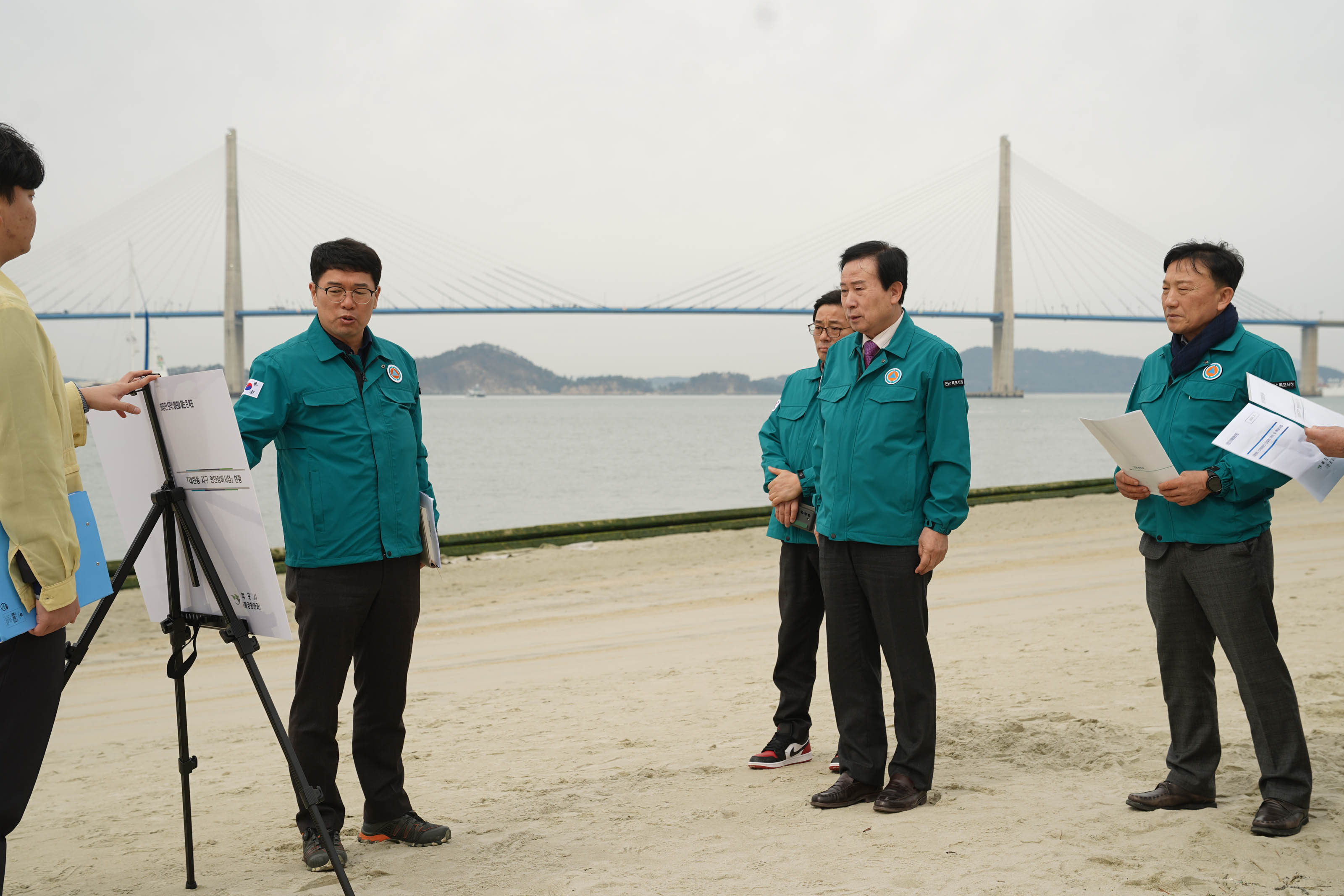 모래로 된 바닥에서 청록색 유니폼을 입은 박홍률 시장과 2명 이상의 직원에게 삭각대에 보드를 세워두고, 무언가를 설명하는 청록색 유니폼을 입은 중년 남성
