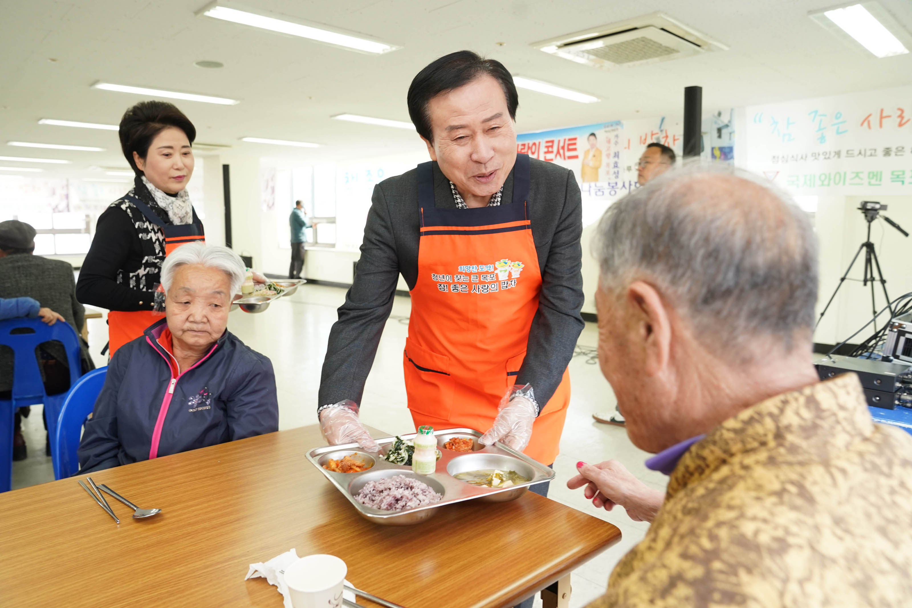 배식받은 식판을 식당 테이블에 앉아계신 두분의 어르신 분들께 직접 전달하고 있는 박홍률 시장의 모습. 웃으며 인사도 건네고 있다. 박홍률 시장 뒤로 중년의 여성이 또 다른 식판을 들고 뒤따라 오고 있다.