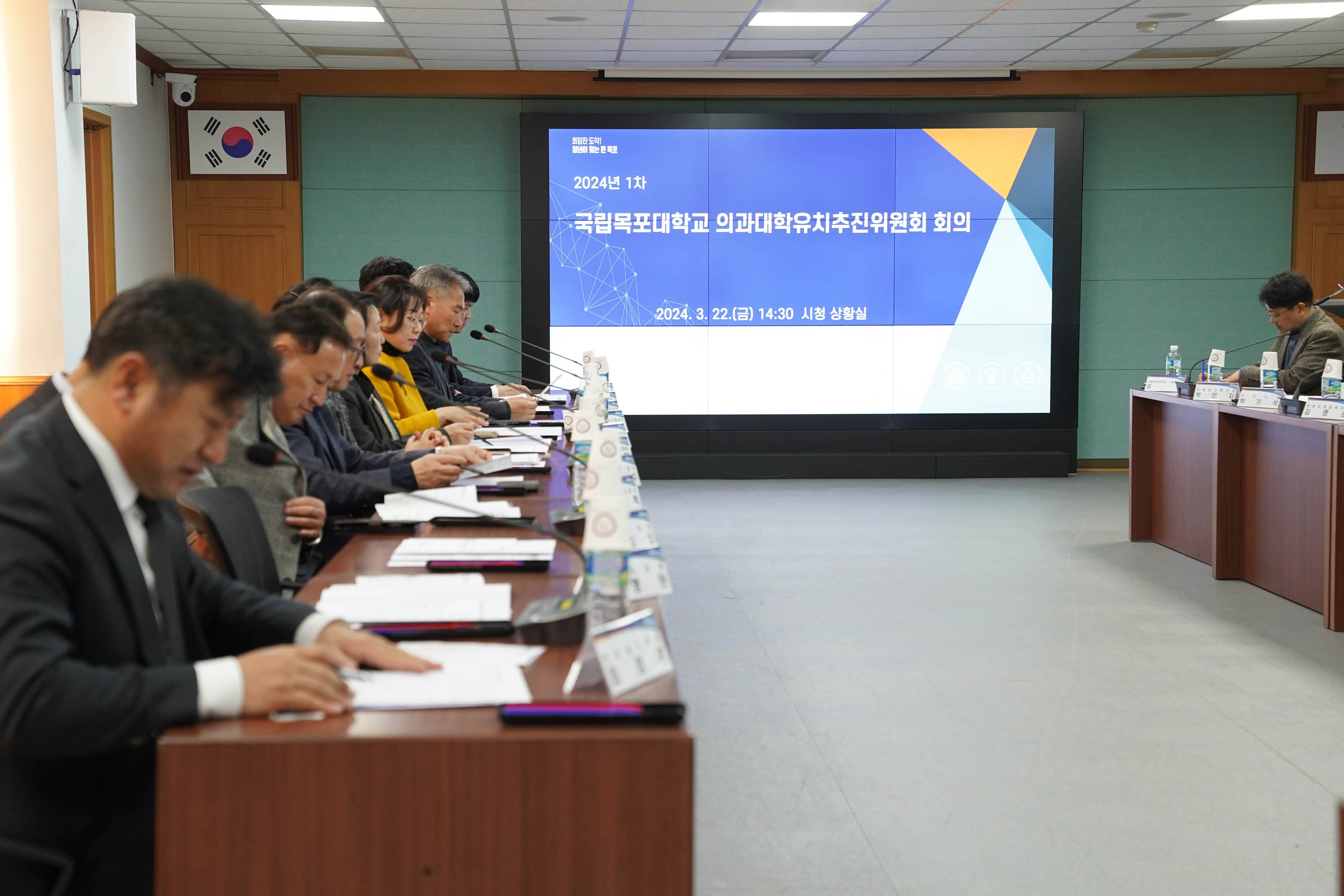 2024년 1차 국립목포대학교 의과대학유치추진위원회 회의 2024.3.22(금) 14:30 시청 상황실 화면이 띄워져 있다. 가로로 긴 회의 테이블에 앉아 있는 위원들. 테이블 위의 종이를 보고 있다.
