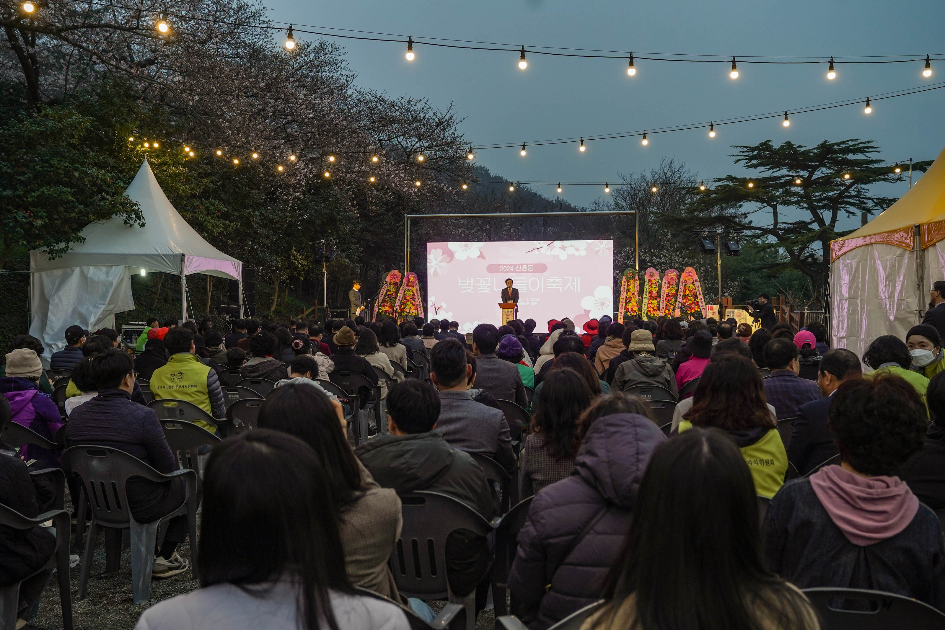 박홍률 시장이 서서 얘기하는 모습이 멀리 보이고, 의자에 앉아 얘기를 듣고 있는 많은 시민들. 어둑해 지는 하늘에 미니 전구들이 빛나고 있다.