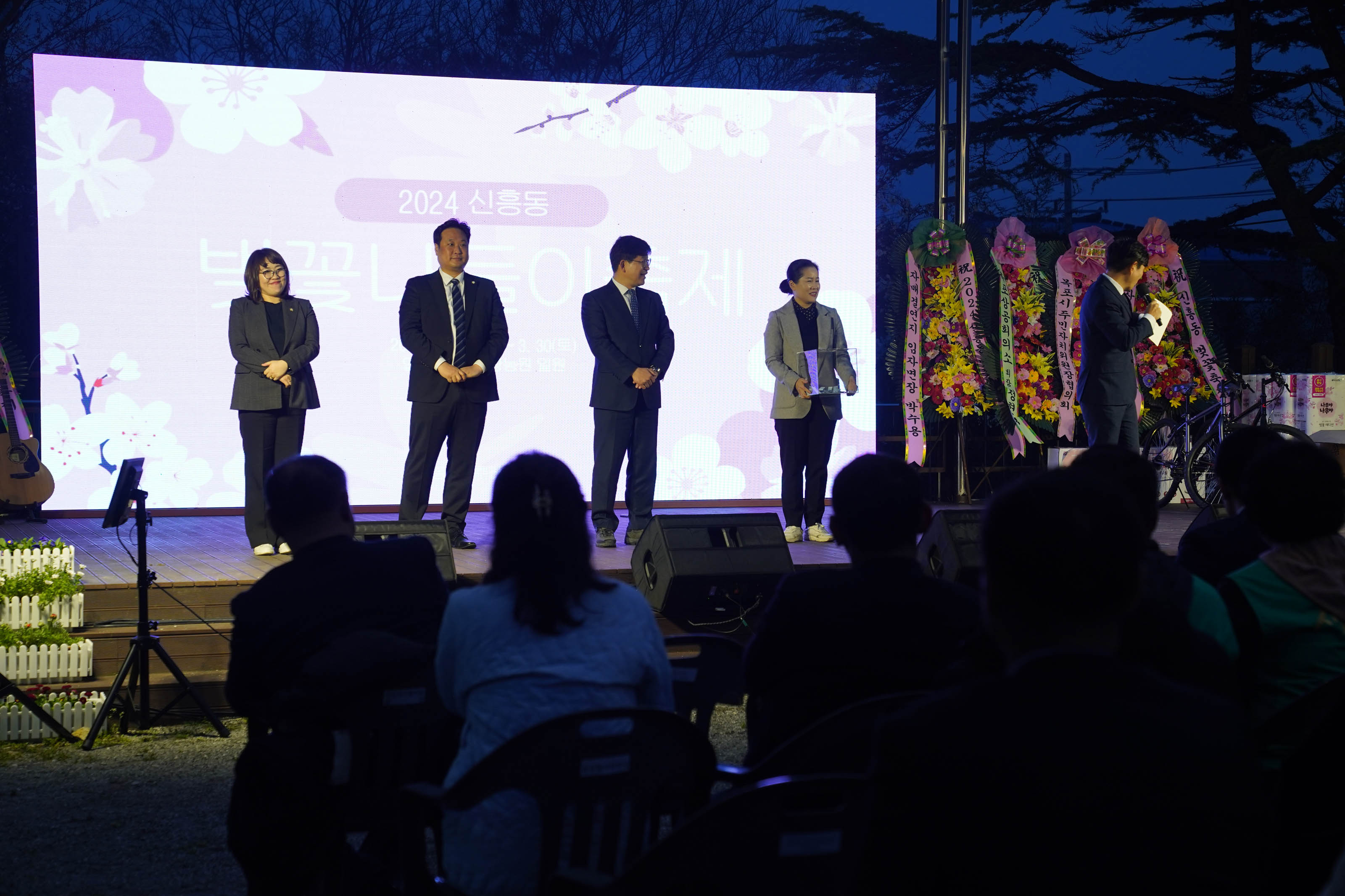 무대 위에 사회자가 마이크를 잡고 이야기하고 있고, 중년 여성 두명, 남성 두명이 여, 남, 남, 여 순으로 서 있다. 4번째 여성은 보라색 띠가 둘러진 투명 상자를 들고 있다.