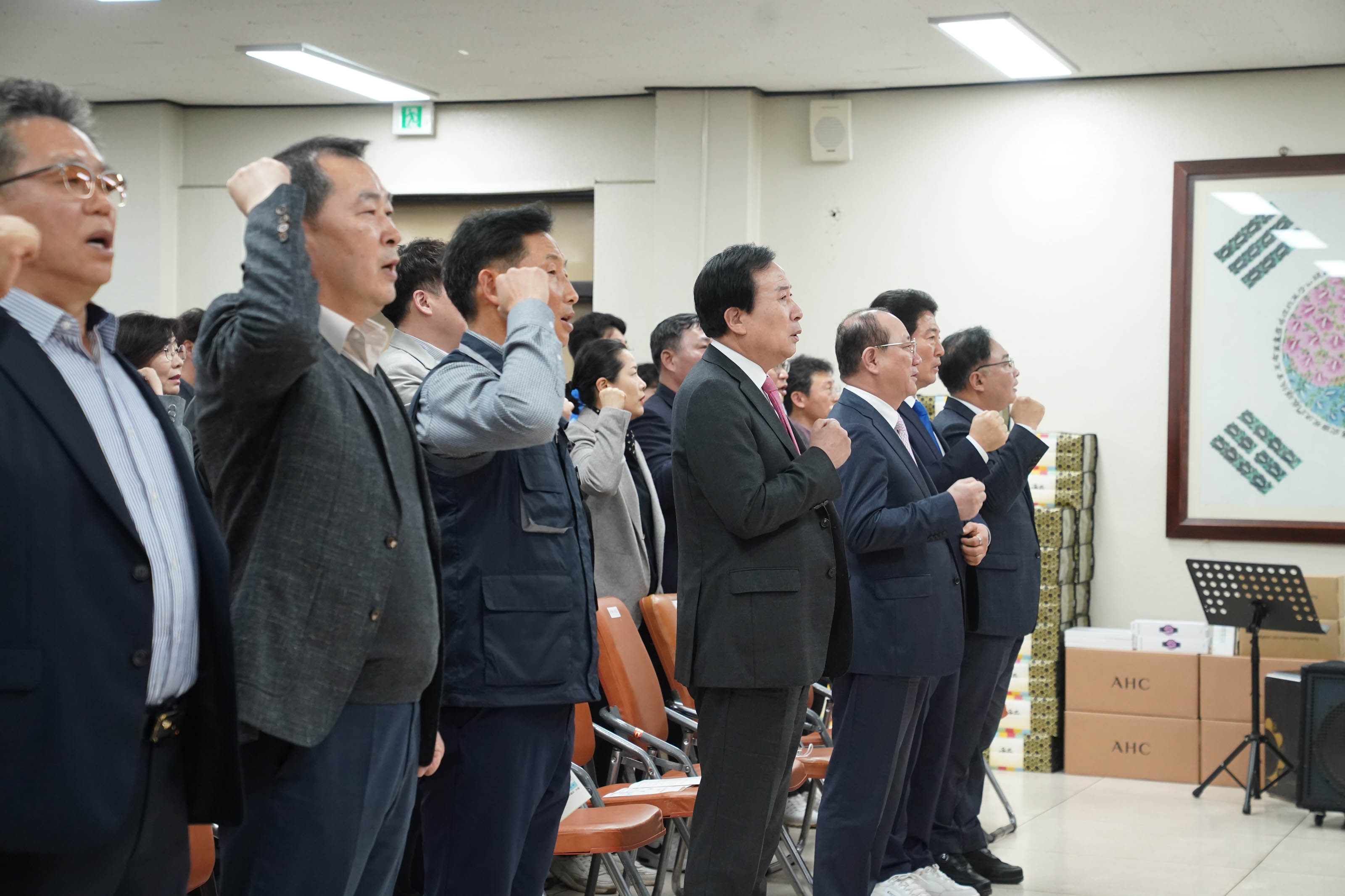 제 11기 목포시공무원노동조합 출범식 2024년 정기총회가 적힌 현수막이 걸려 있고, 노동조합 조끼를 입은 사람들이 나란히 서있다. 그 앞에 다표로 보이는 중년의 남성이 목포시공무원노동조합이 적힌 하얀색에 노란색 술이 달린 커다란 깃발을 흔들고 있다.