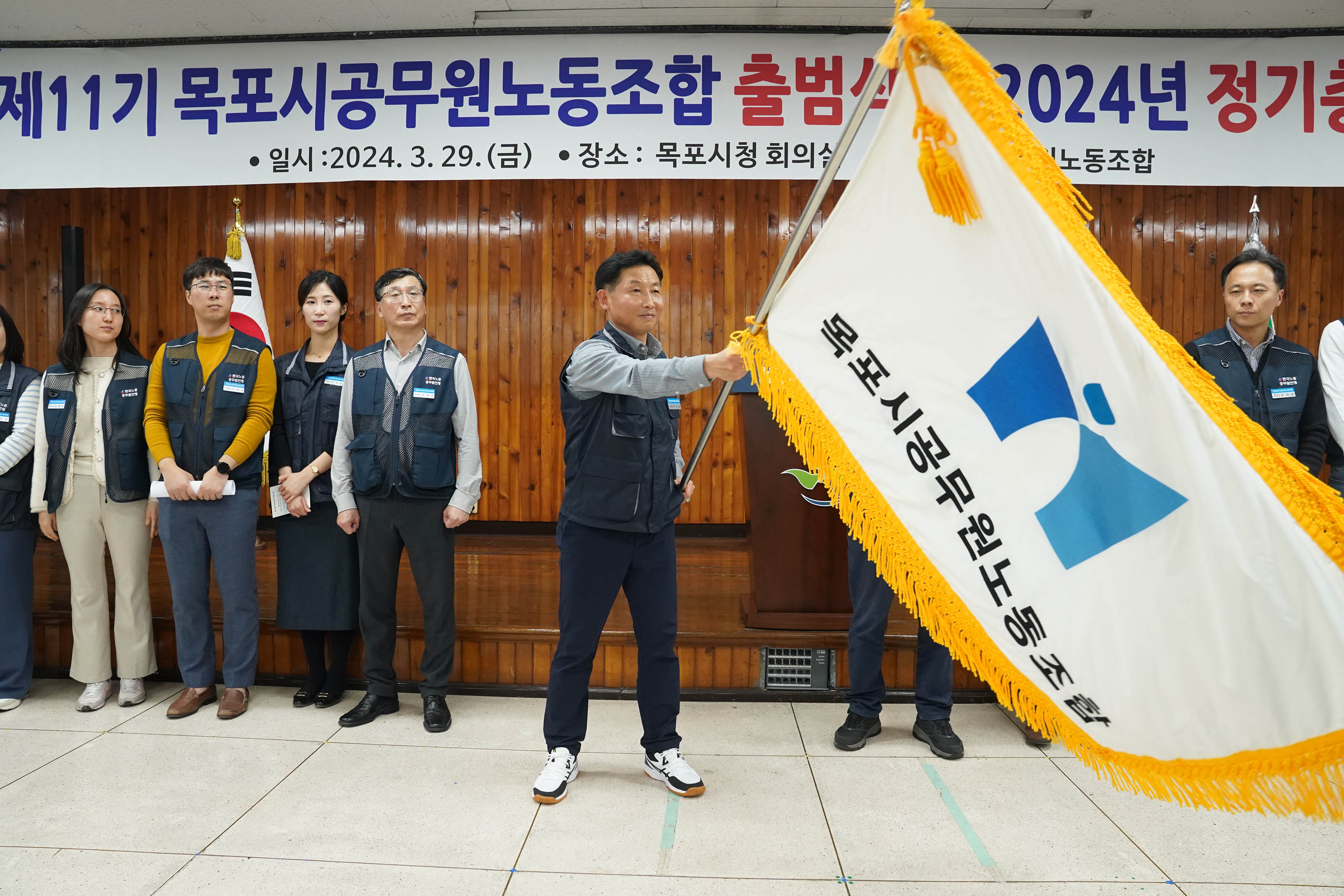제 11기 목포시공무원노동조합 출범식 2024년 정기총회가 적힌 현수막 앞에서 회색 니트와 회색 자켓을 입고 꽃다발과 명패 비슷한 사진을 들고 있는 중년 남성의 양쪽으로 노동 조합 조끼를 입은 남성과 베이지색 코트를 입고 안경을 낀 남성이 서서 찍은 사진