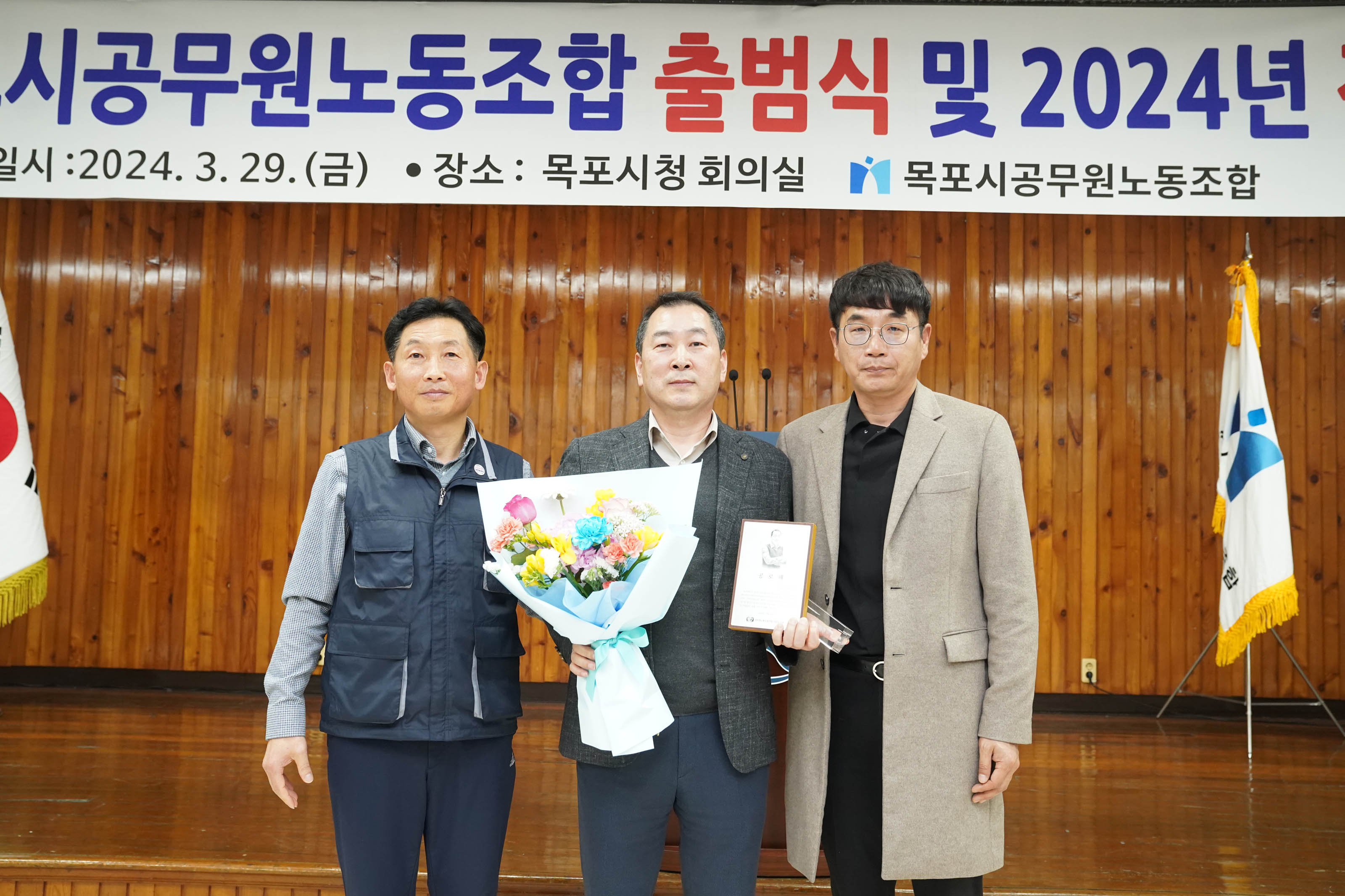 제 11기 목포시공무원노동조합 출범식 2024년 정기총회가 적힌 현수막이 걸려 있고, 희망찬 도약! 청년이 찾는 큰 목포 문구가 적힌 단상에서 꽃다발을 들었던 남성이 얘기하고 있고 양 옆으로 태극기과 노동 조합 깃발이 세워져있다. 그 앞에 앉아 얘기를 듣고 있는 사람들의 사진