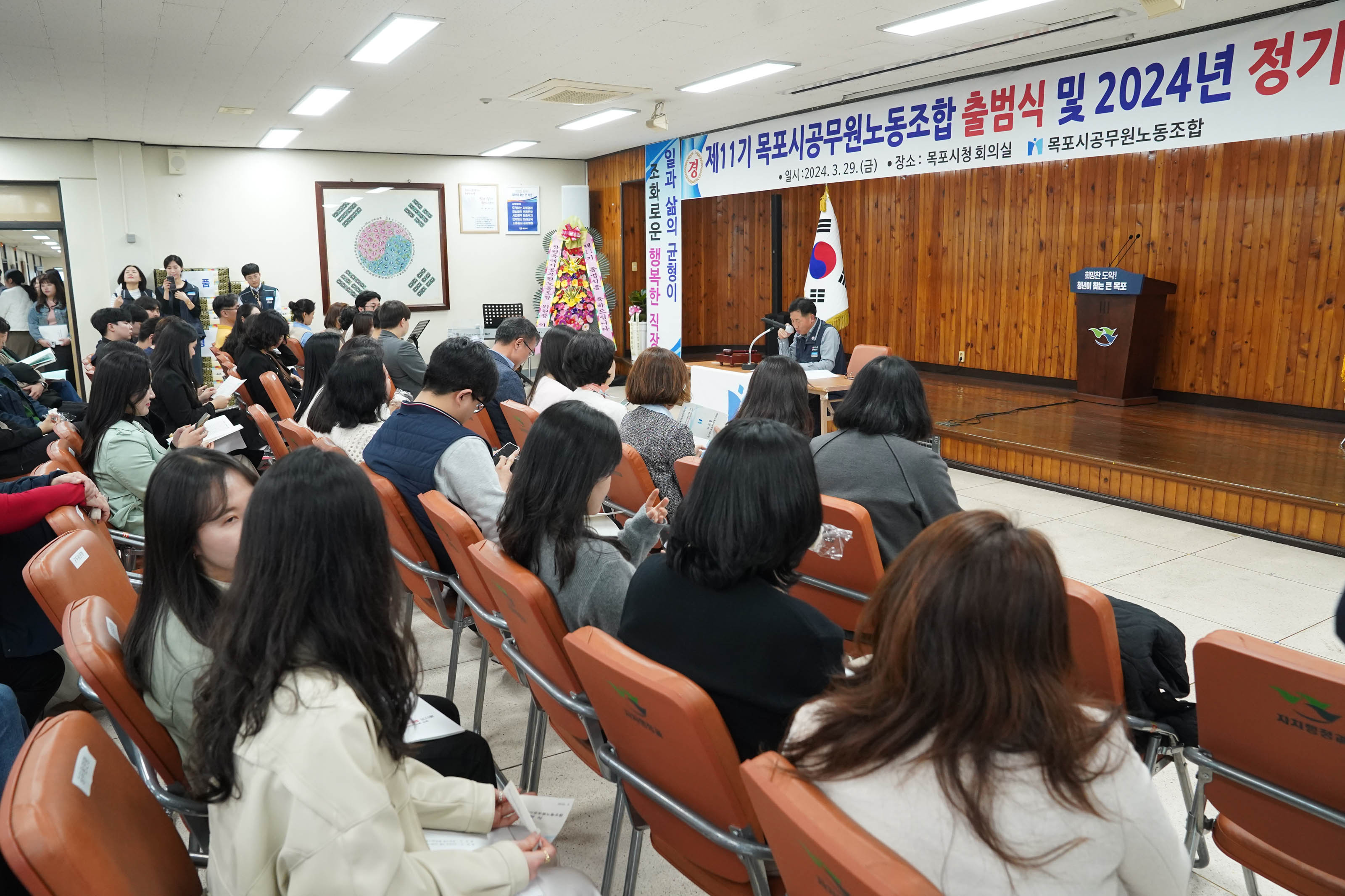 제 11기 목포시공무원노동조합 출범식 및 2024년 정기총회 일시:2024.3.29(금) 장소:목포시청 회의실 목포시공무원노동조합 현수막 아래로 앉아서 글을 낭독하는 중년의 남성 얘기를 듣고 있는 공무원들이 앉아있다.